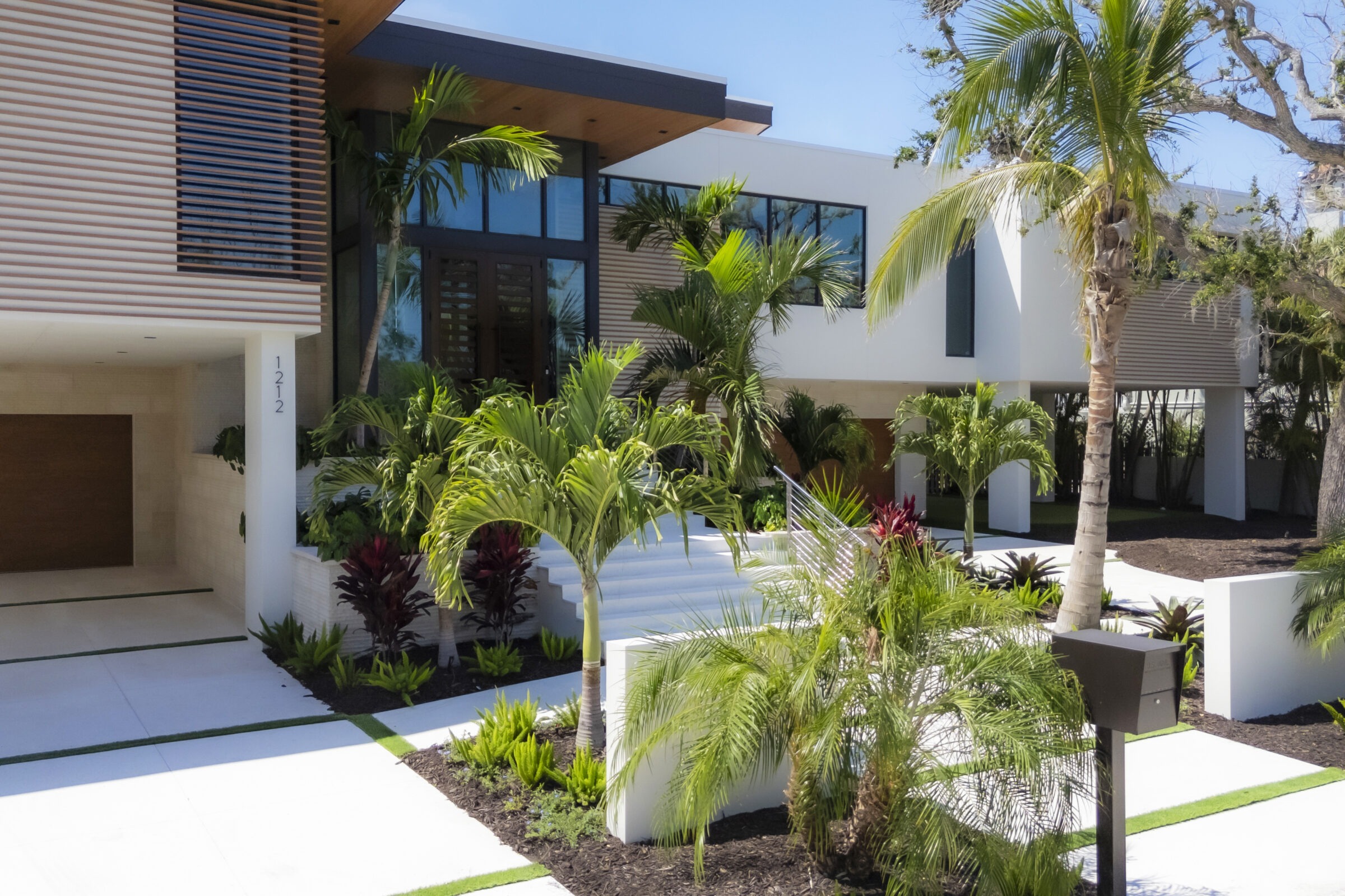 Modern house with large windows, surrounded by tropical plants and palm trees. Sleek design features horizontal wooden slats, white walls, and a garage.