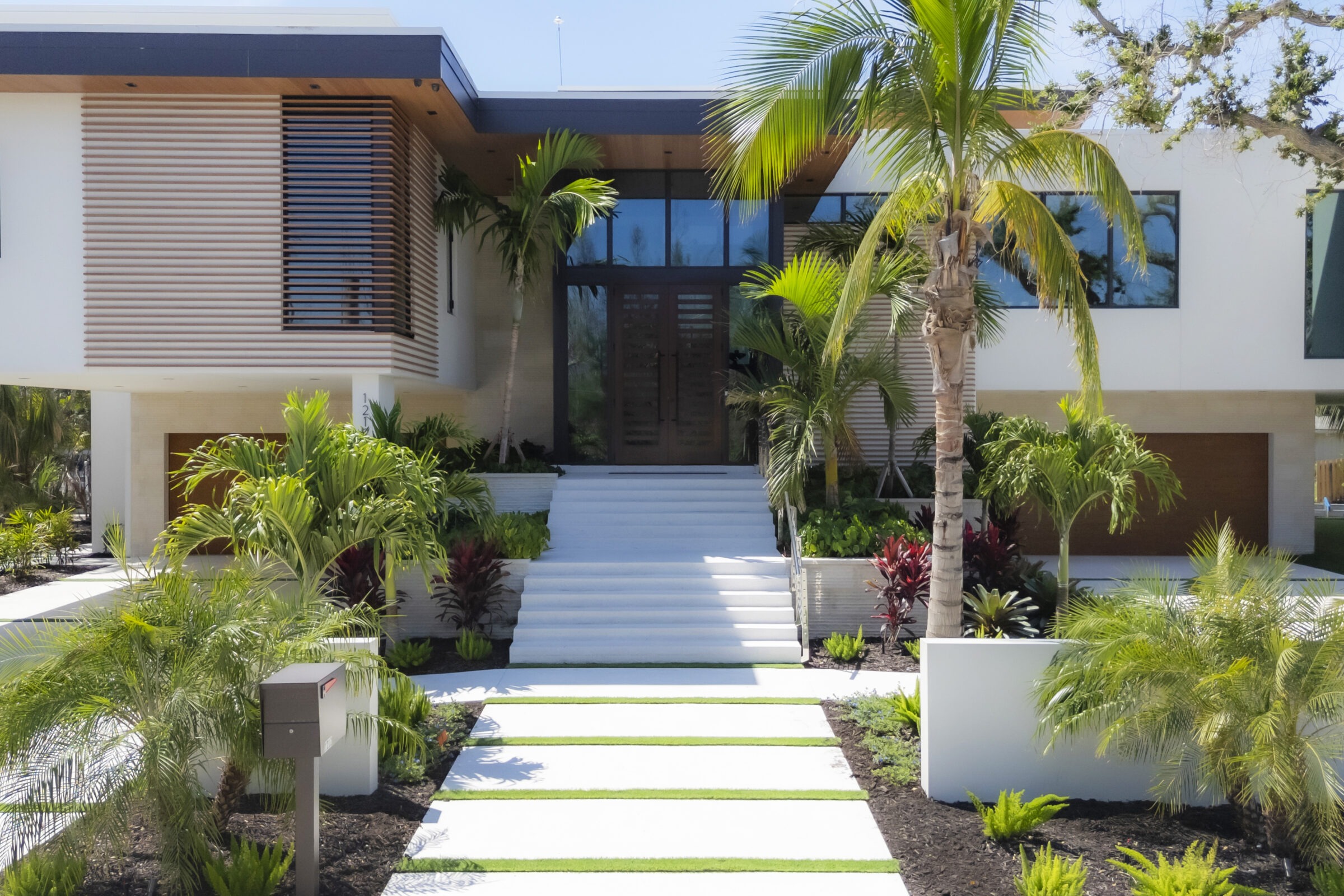 Modern house with tropical landscaping, featuring palm trees, wide steps, and large glass windows. A concrete path leads to the entrance.