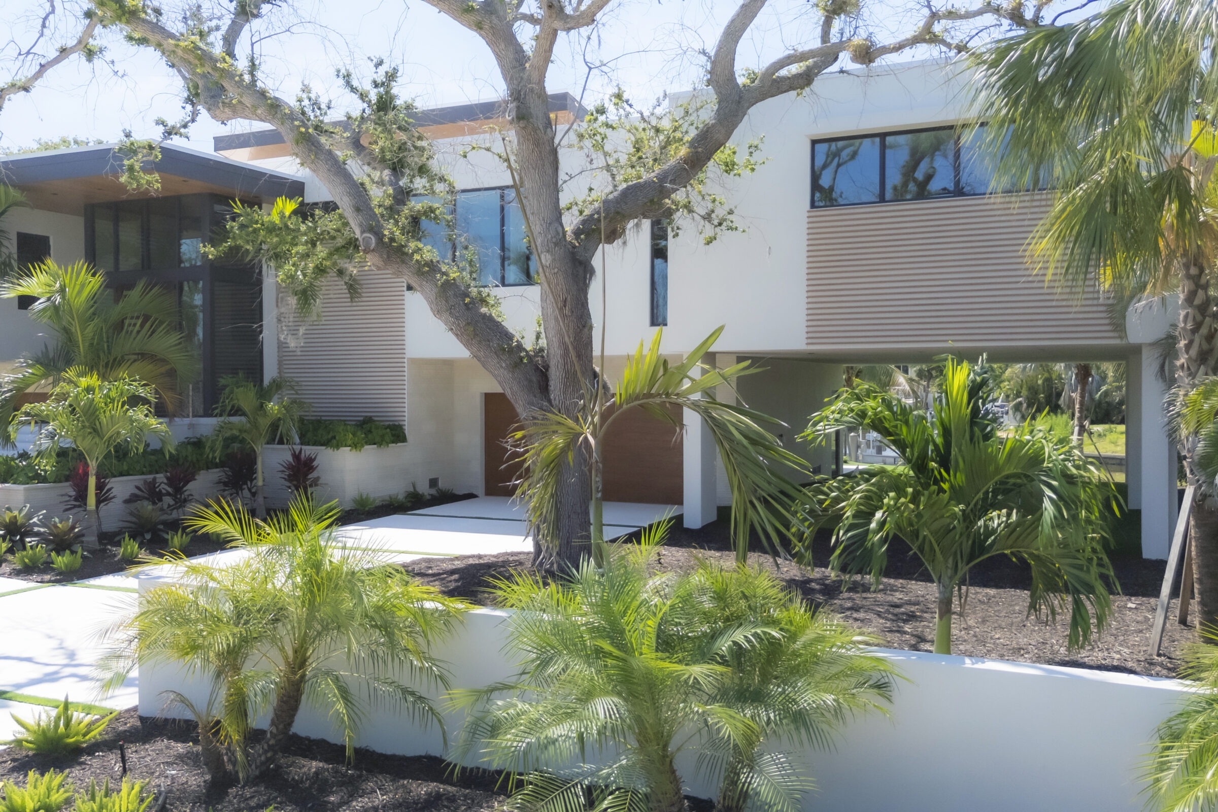 Modern house with large windows surrounded by tropical greenery, trees, and landscaped gardens, creating a serene and inviting atmosphere in a sunny setting.