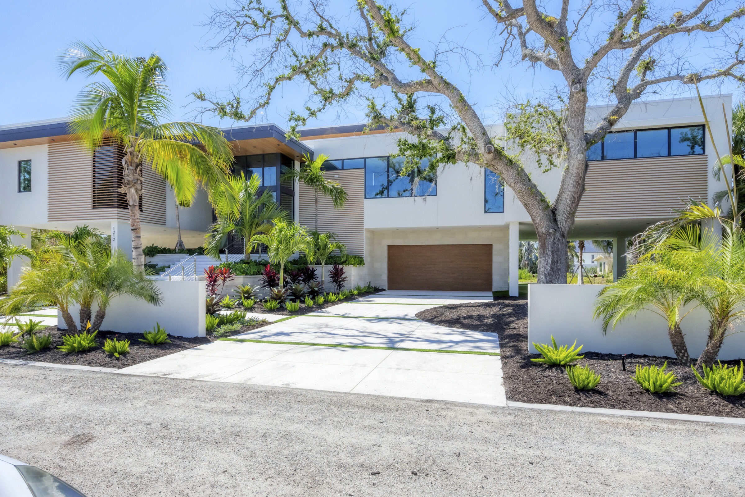 Modern house surrounded by tropical plants, trees, and shrubs. Features large windows and wooden accents, with a driveway leading to a two-car garage.