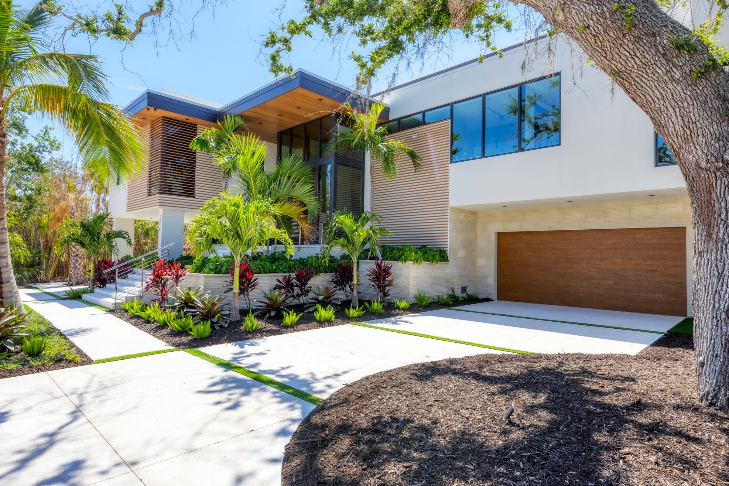 Modern house with large windows, wooden accents, surrounded by lush green plants and palm trees, featuring a two-car garage and sleek driveway.