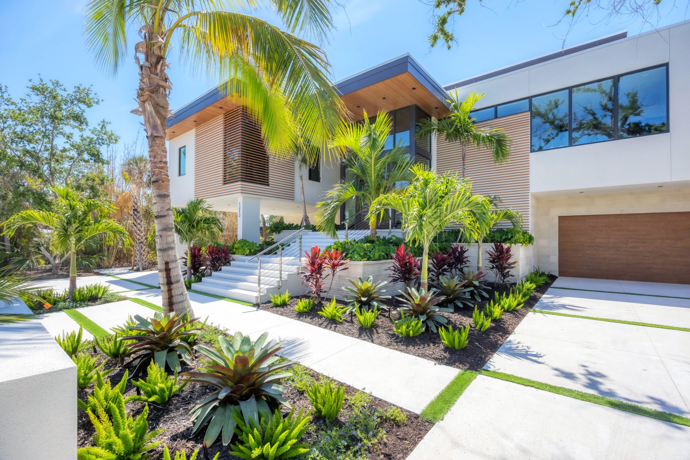 Modern house with palm trees and lush landscaping. Clean lines, wooden accents, large windows, and a spacious driveway create a sleek, tropical appearance.