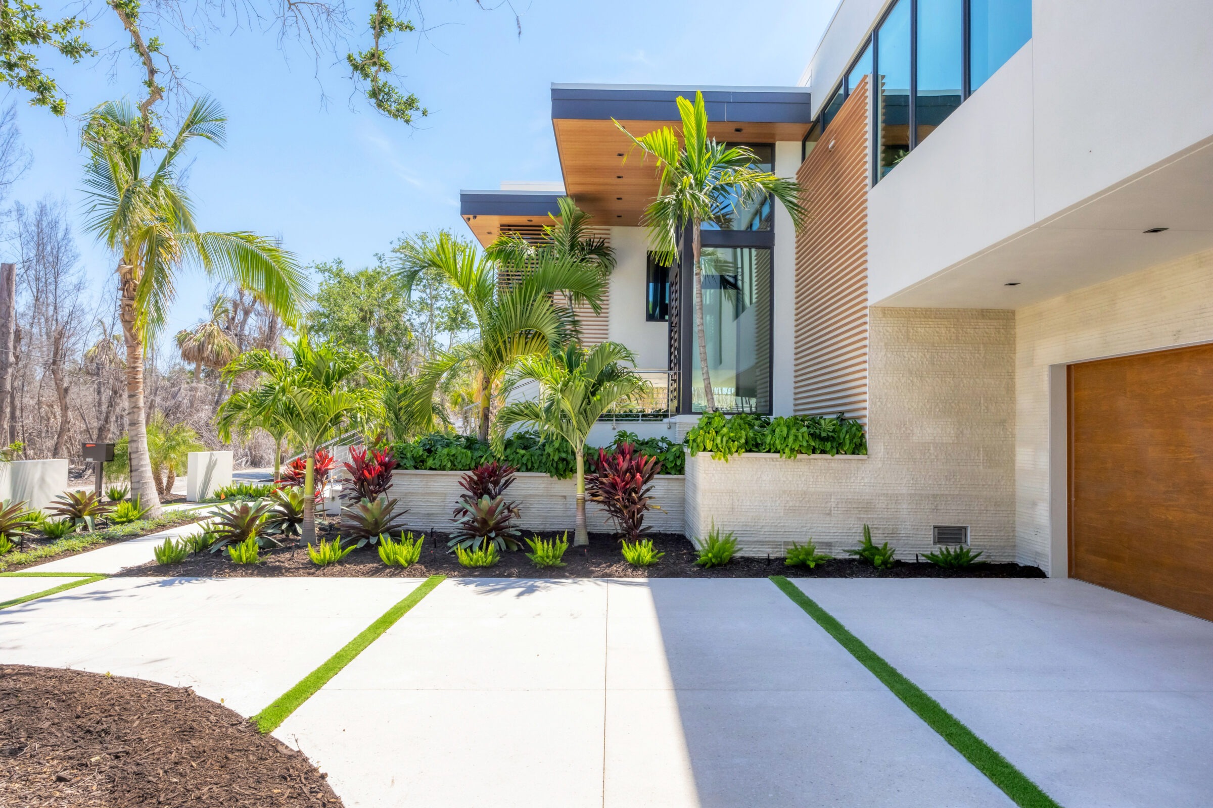 Modern house with large windows, lush tropical landscaping, and a spacious driveway. Palm trees and vibrant greenery enhance the contemporary design.