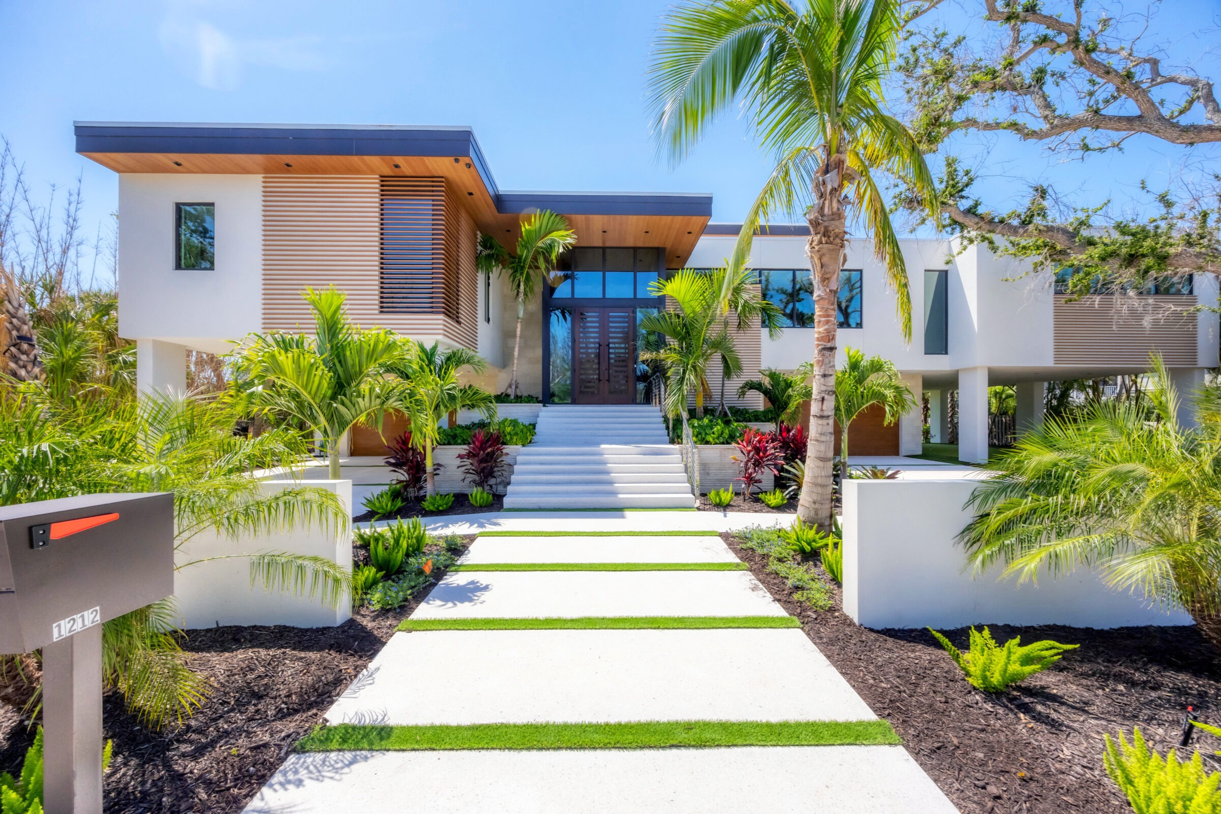 Modern house with palm trees, landscaped garden, and wide steps. A sleek mailbox sits near the entrance. Bright, sunny day setting.
