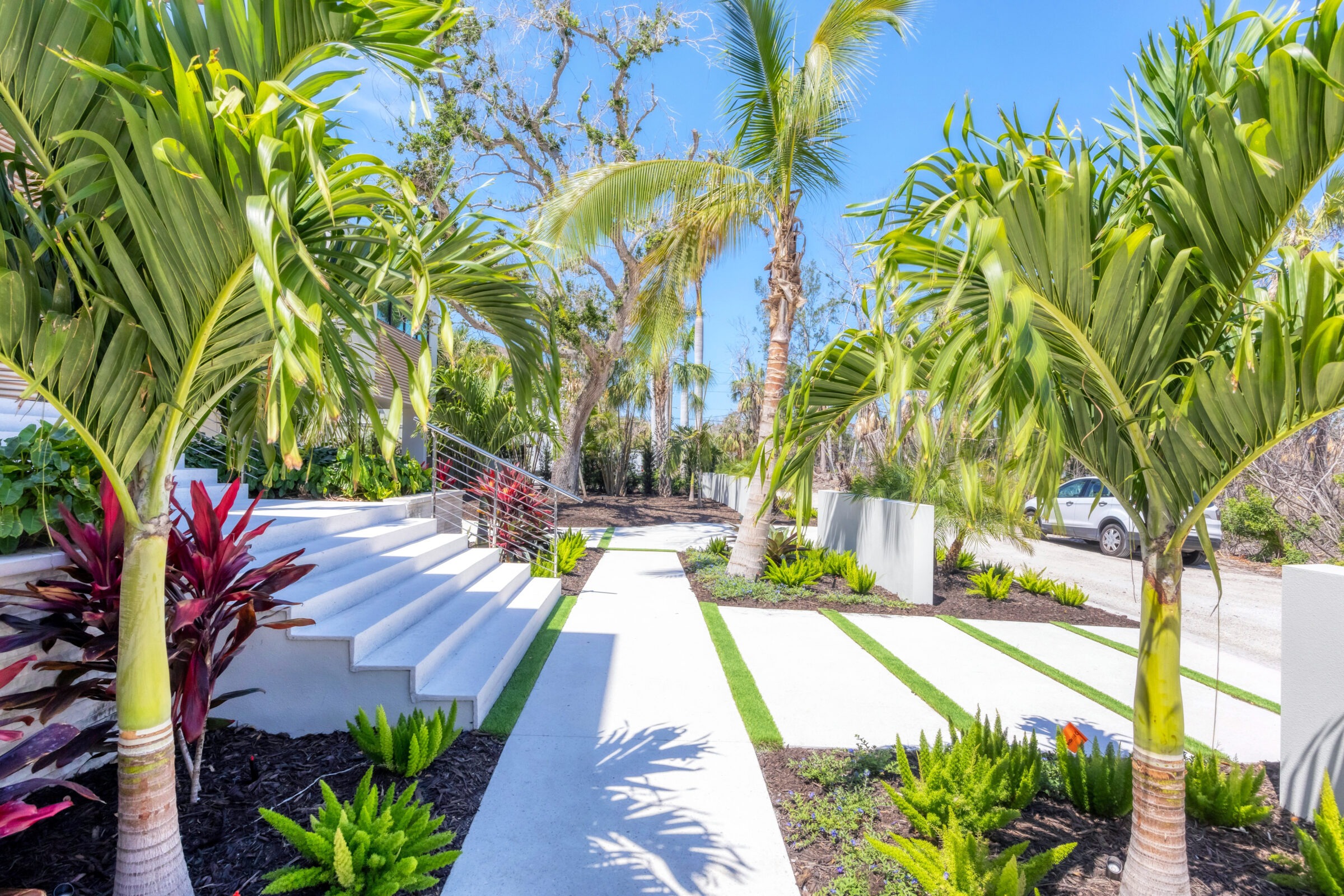 Tropical garden entrance with palm trees, vibrant foliage, modern steps, and a walkway leading to a parked white vehicle under a clear blue sky.