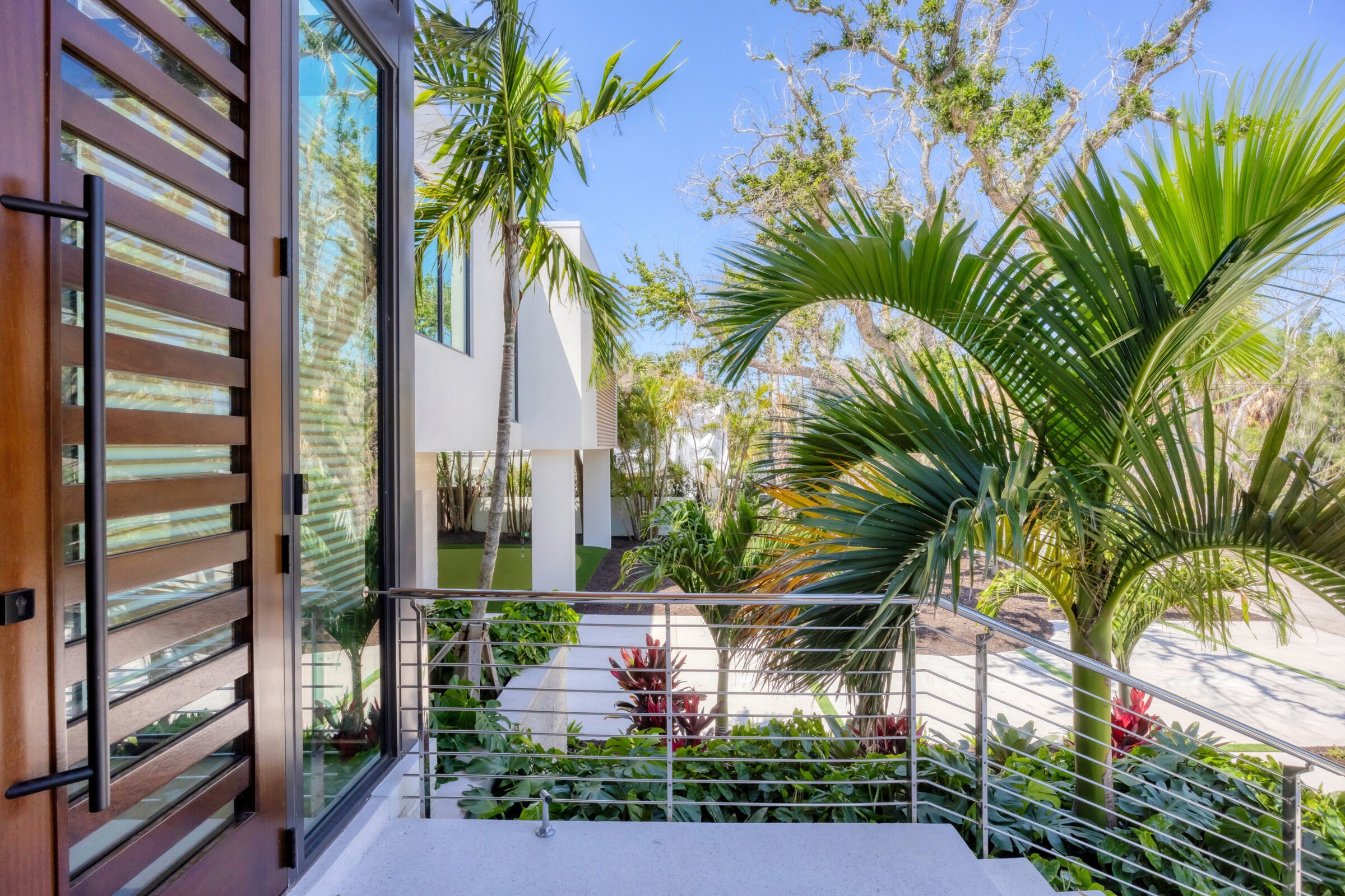 Modern house entrance with wooden door, glass panels, and lush tropical plants. Sunny, clear blue sky enhances the vibrant greenery.