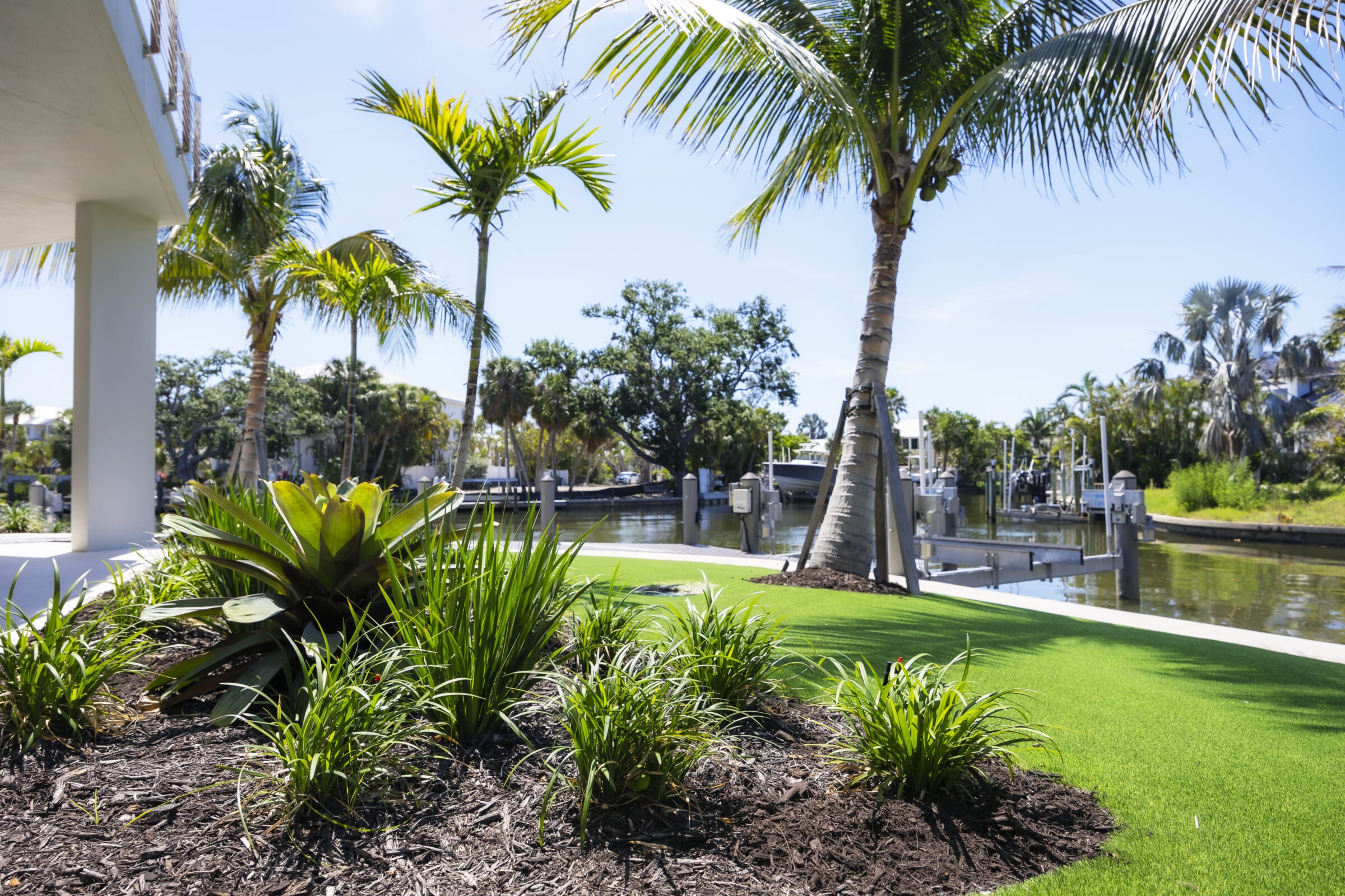 Lush greenery and palm trees frame a serene canal with boats docked, under a clear blue sky, beside a modern structure.