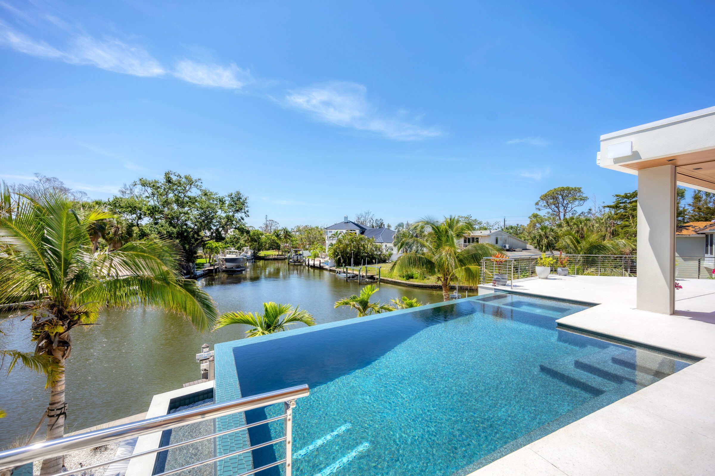 Infinity pool overlooking a scenic canal surrounded by palm trees and modern houses under a clear blue sky. Peaceful and luxurious atmosphere.