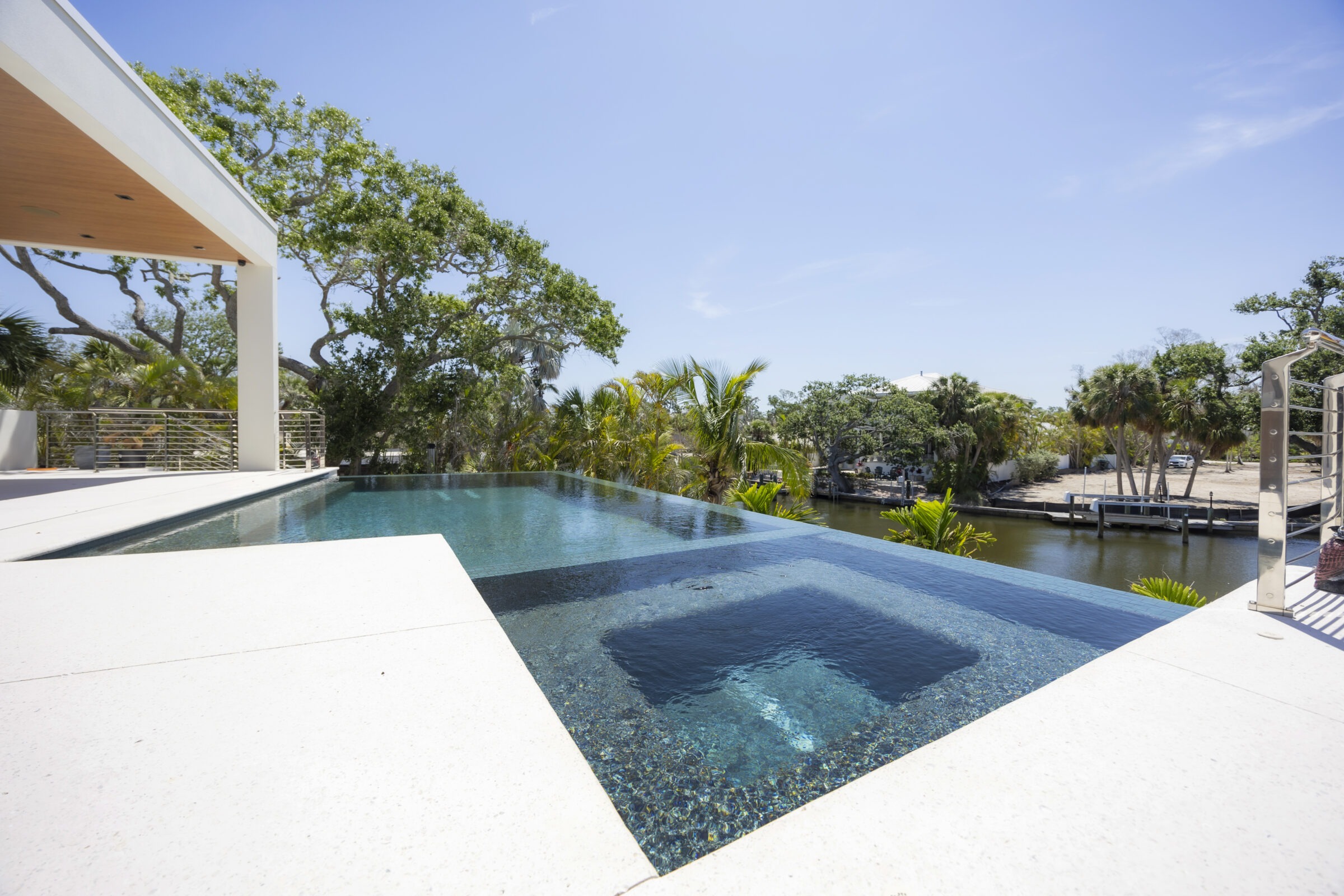 Infinity pool overlooking a serene canal, surrounded by lush greenery and modern architecture. Clear blue sky enhances the tranquil, luxurious setting.