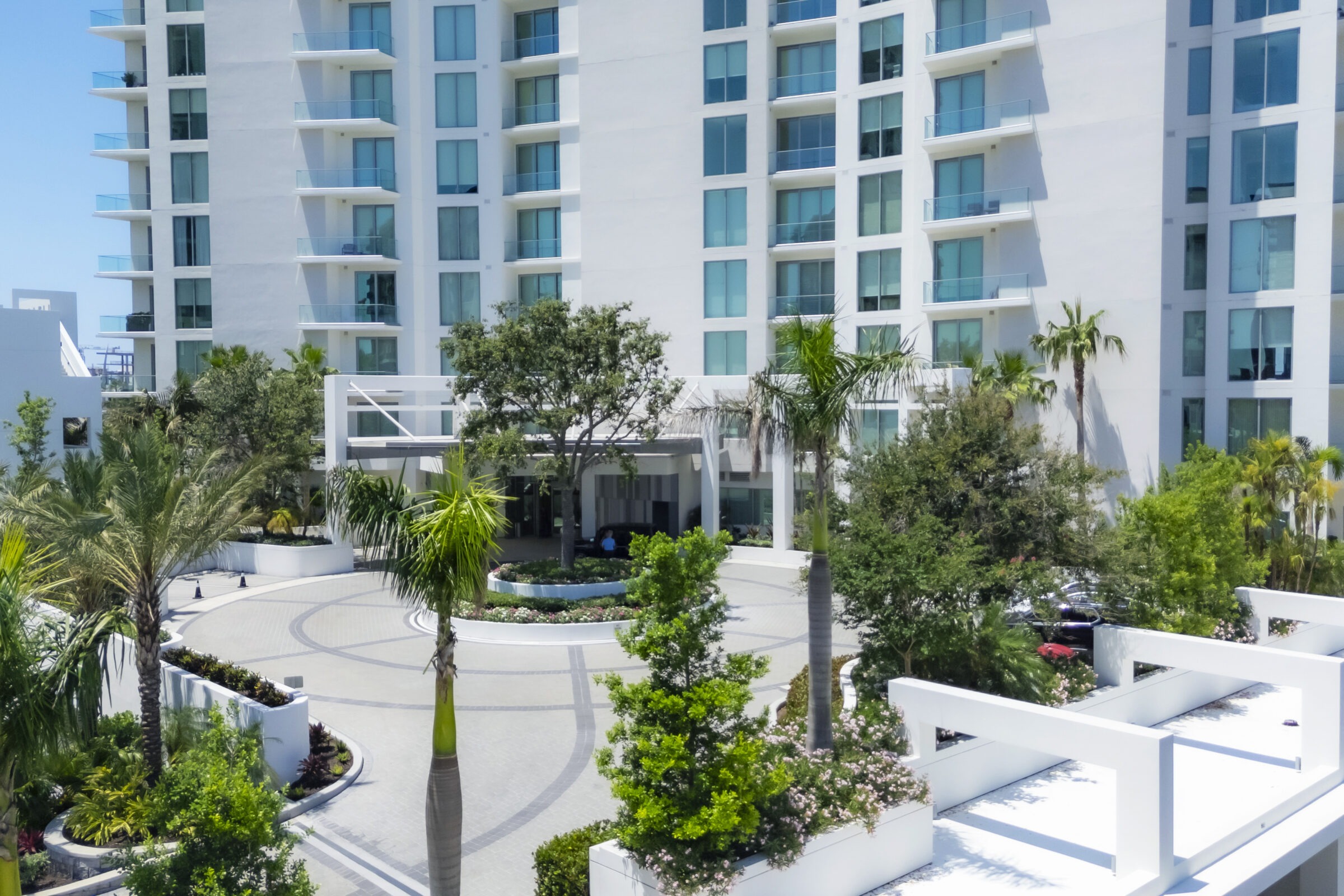Modern white building with balconies, surrounded by lush green palm trees and landscaped gardens. Person seated under tree near main entrance.