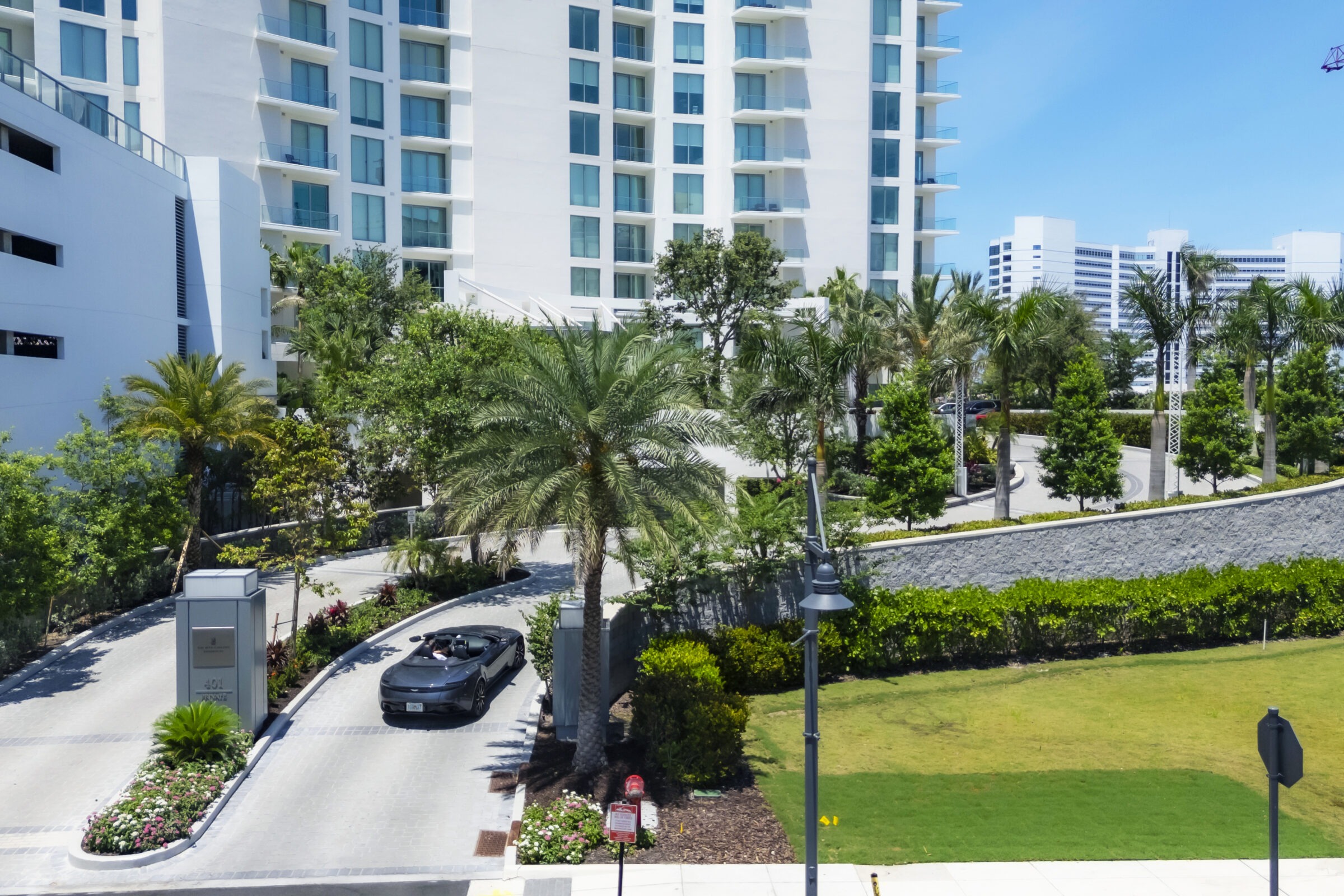 A convertible car drives through a landscaped urban area with tall buildings and palm trees under a clear blue sky.