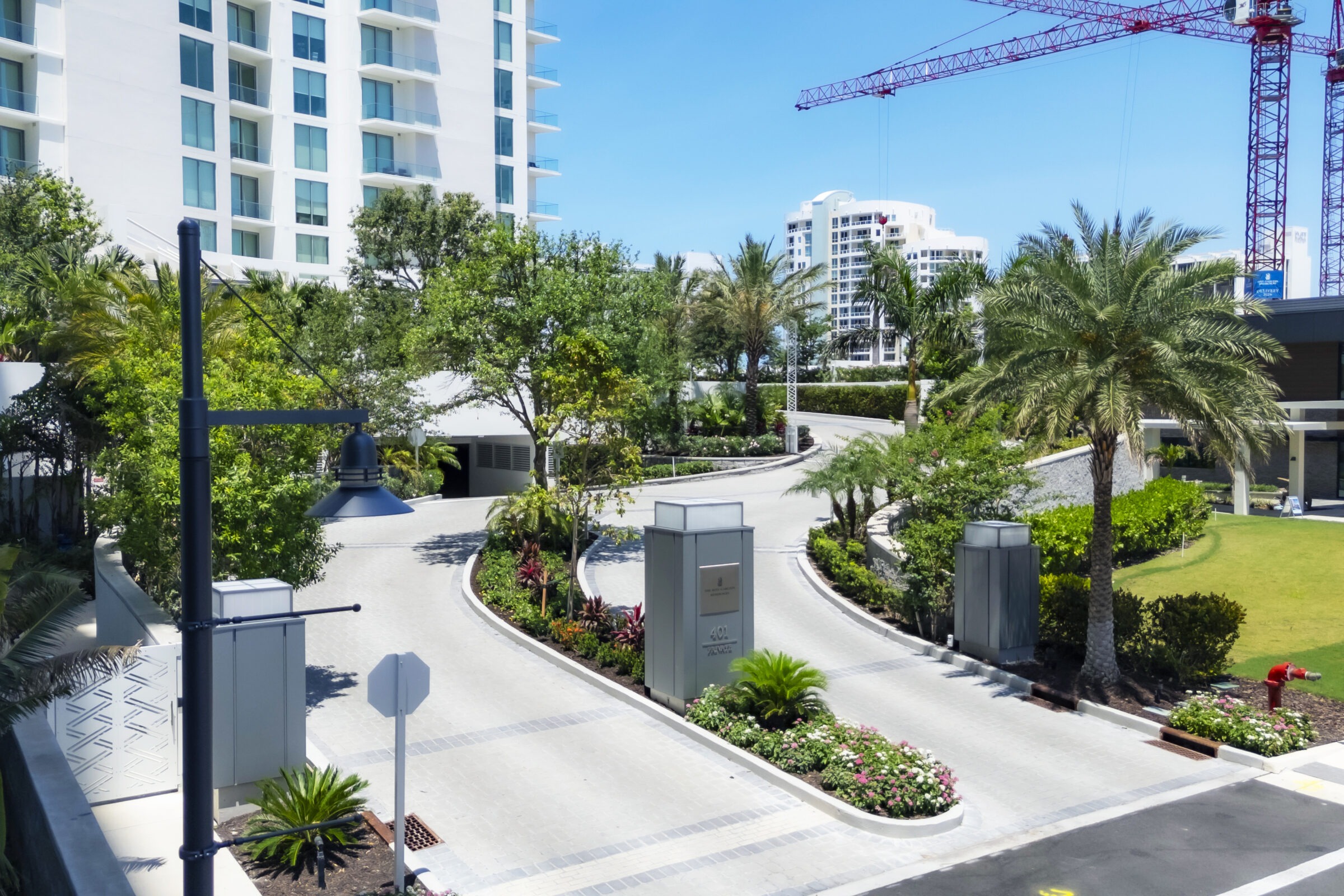 Modern residential complex with lush gardens, palm trees, and construction cranes in the background. A curving driveway leads to a garage entrance.