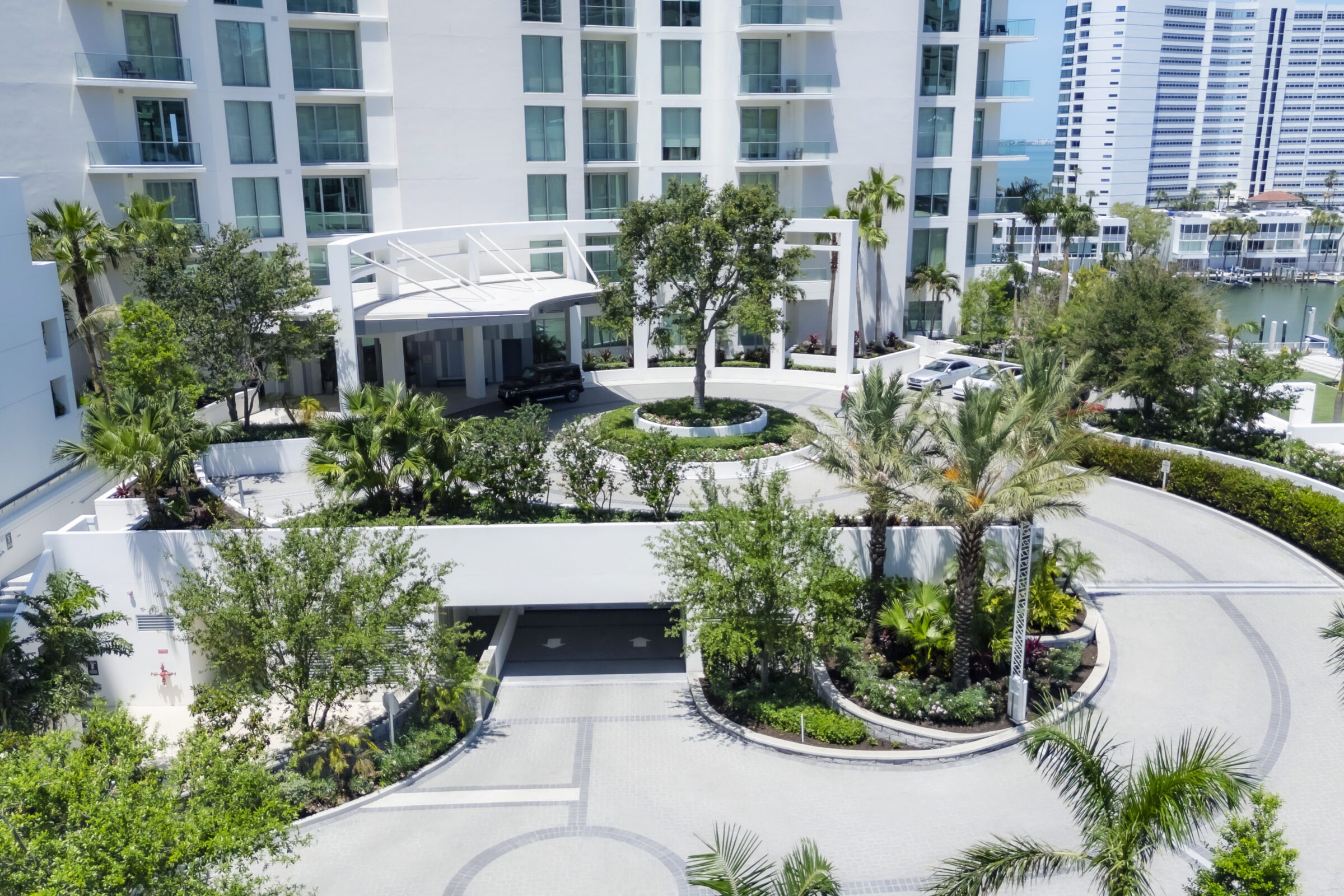 Modern white building entrance with a landscaped circular driveway, surrounded by lush greenery and palm trees, adjacent to residential waterfront properties.