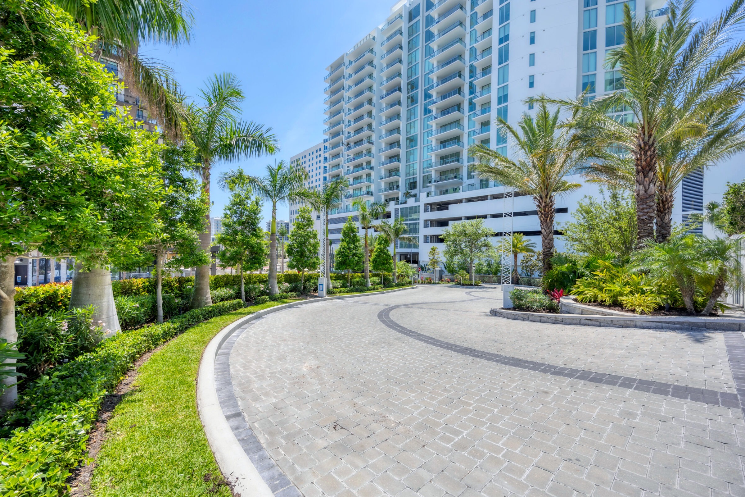 A modern high-rise building surrounded by lush palm trees, landscaped gardens, and a curved stone pathway under a clear blue sky.