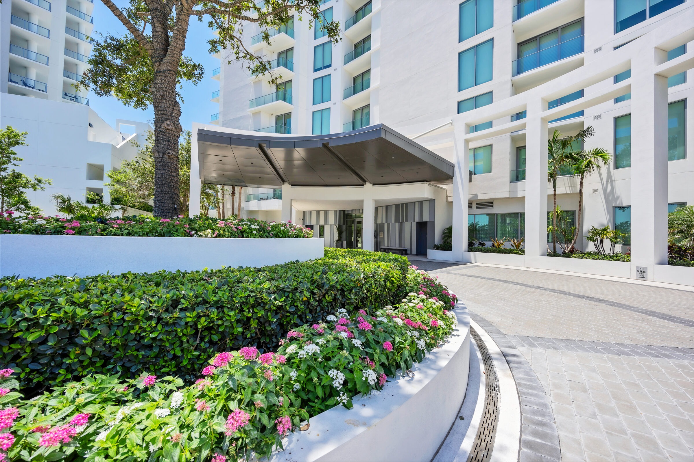 Modern building entrance features lush landscaping with colorful flowers and palm trees, under a bright blue sky. No people are visible.