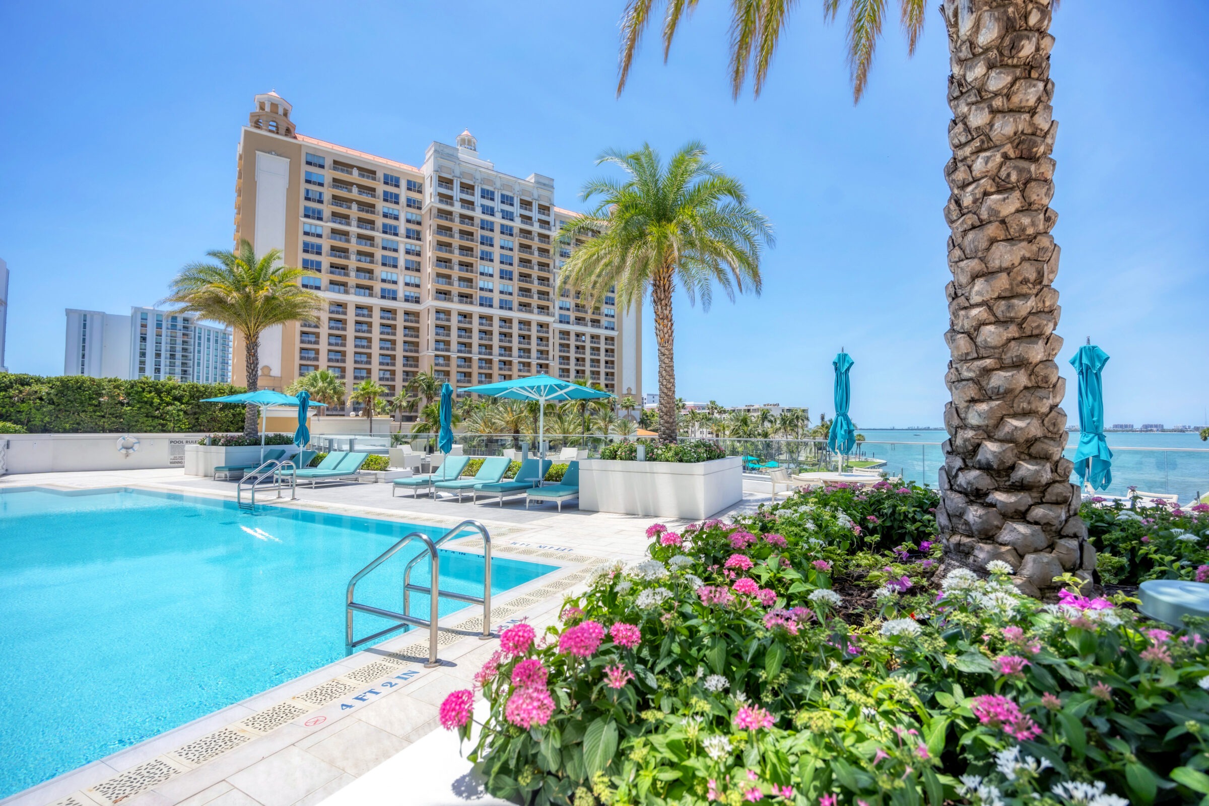 A luxurious poolside scene with lounge chairs, umbrella, flowers, palm trees, and a tall hotel building by a serene blue ocean.