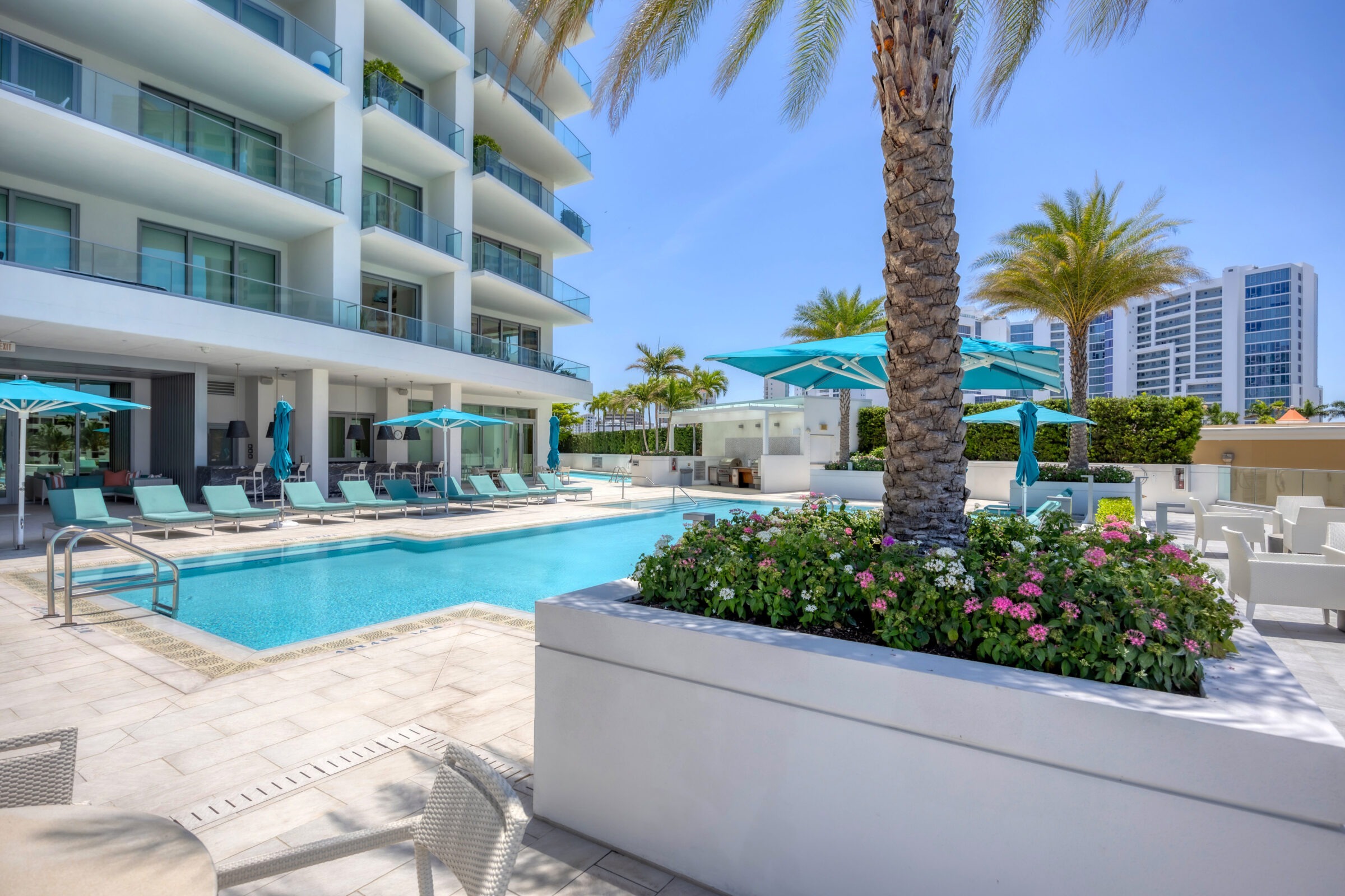 Modern hotel pool area with turquoise lounge chairs and umbrellas, surrounded by palm trees and flowers. High-rise buildings visible in the background.