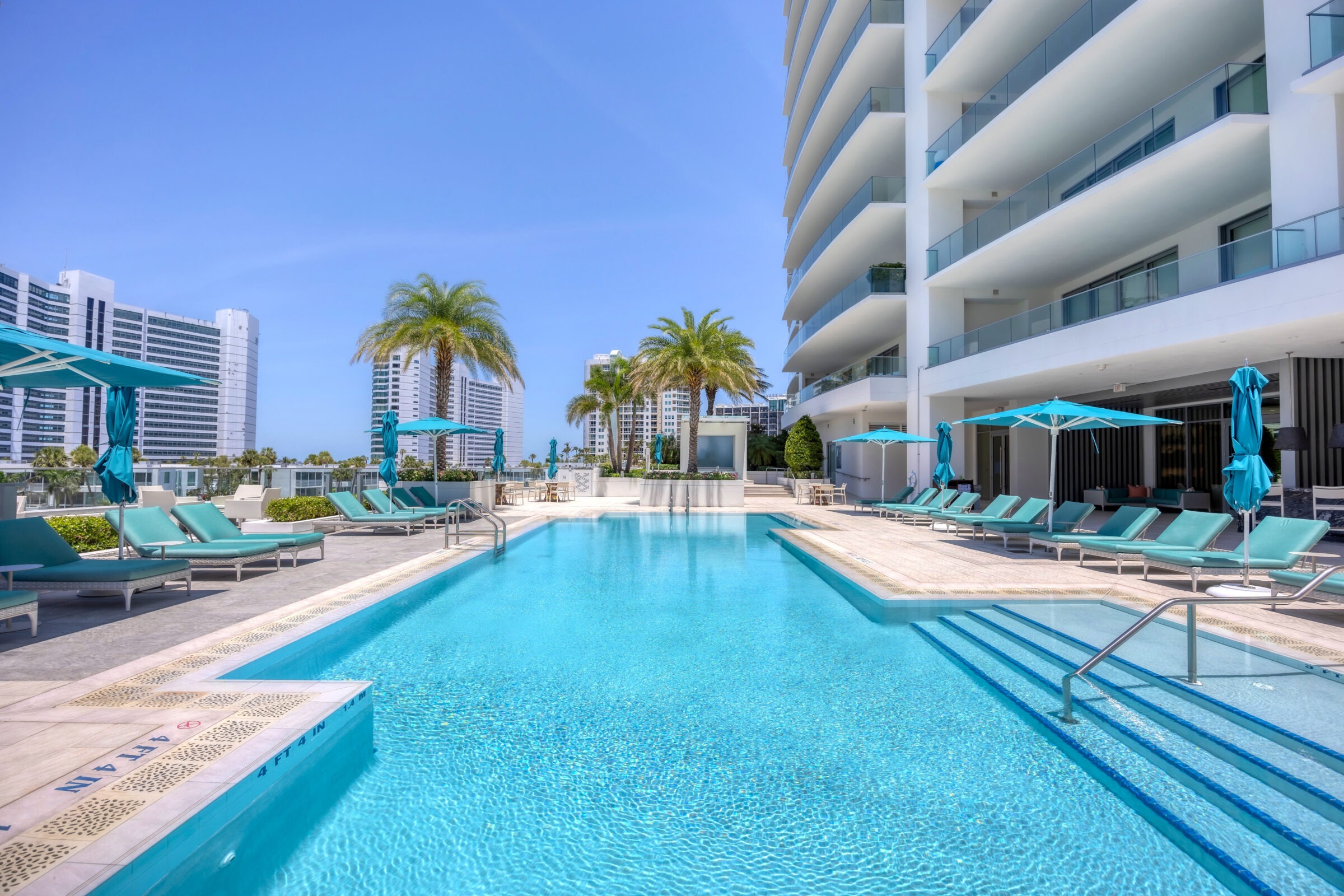 Modern skyscrapers surround a peaceful rooftop pool with turquoise lounge chairs and umbrellas under a clear blue sky. No people present.