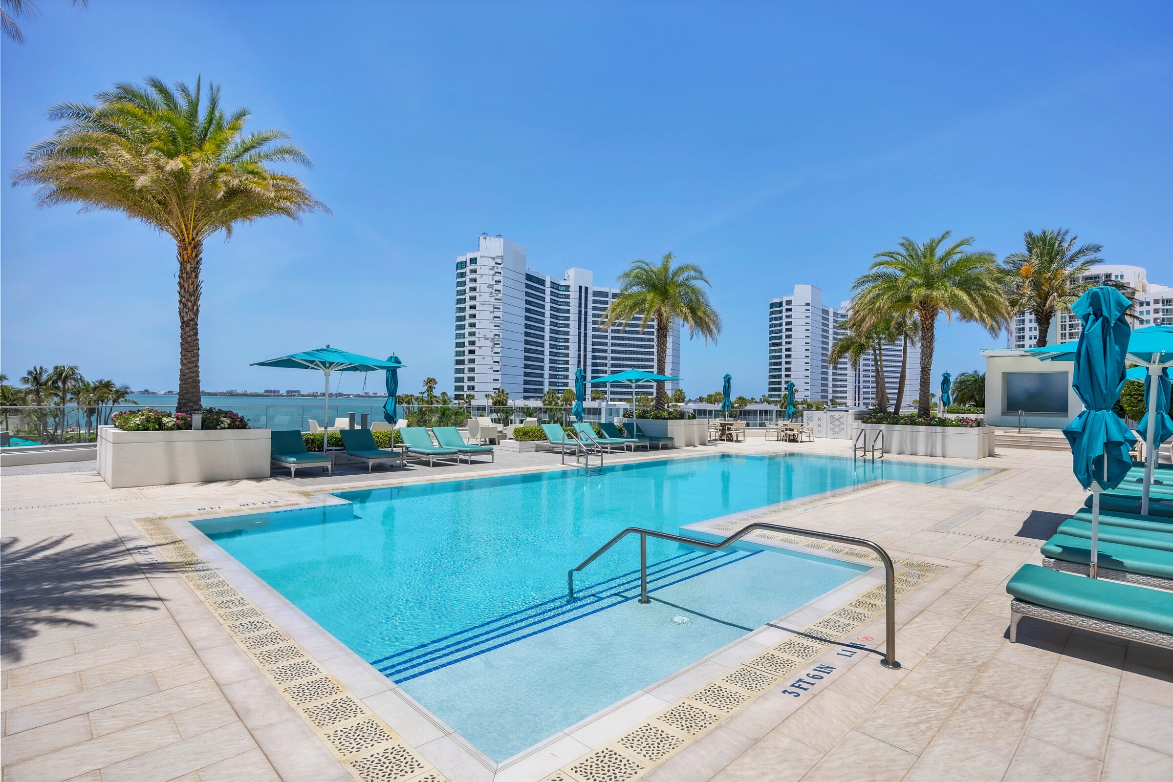 Modern outdoor swimming pool with turquoise loungers and umbrellas, surrounded by palm trees. Skyscrapers and blue sky in the background.
