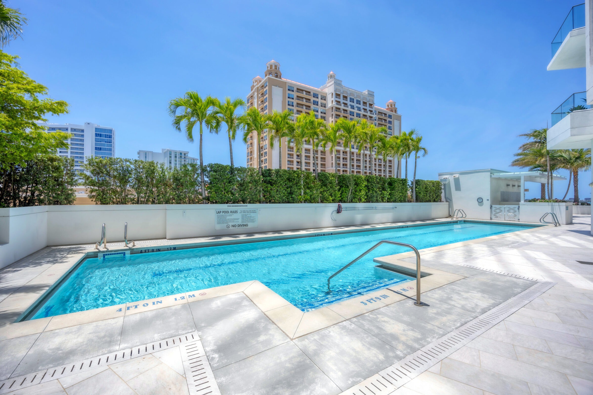 A clear pool on a sunny day, surrounded by palm trees, with a tall building in the background.