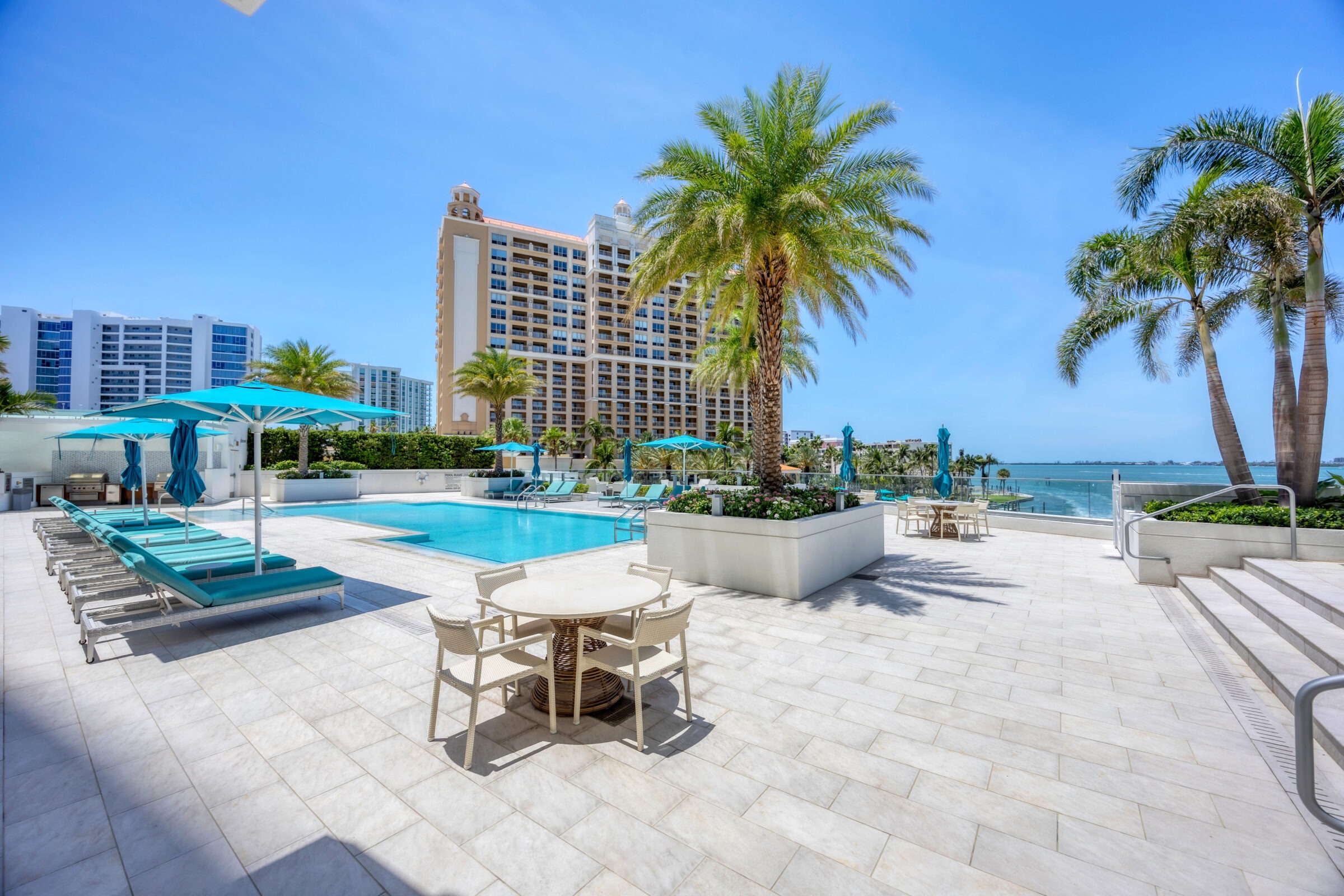 Modern poolside area with palm trees, turquoise umbrellas, lounge chairs, and tables. Background features high-rise buildings by the waterfront under a clear sky.