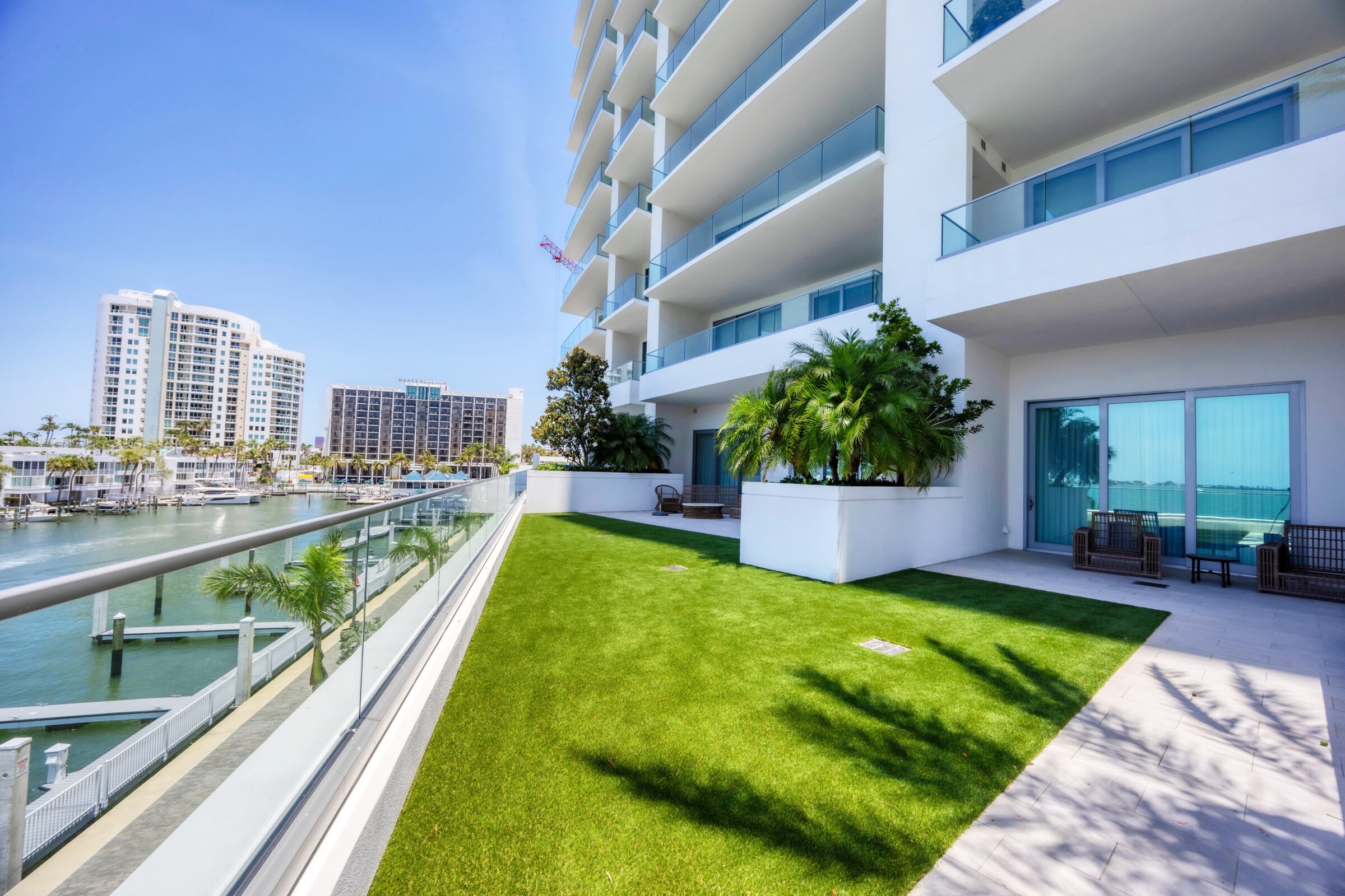 Modern waterfront apartment with lush lawn, overlooking calm marina. Tall buildings and clear sky in the background. Luxurious, open, and serene setting.