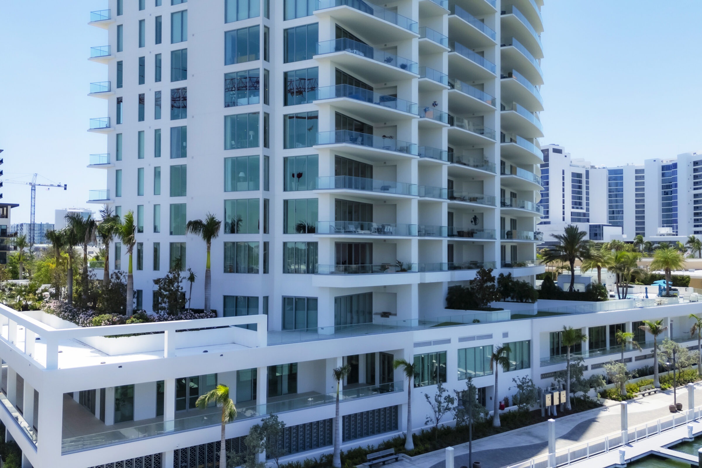Modern multi-story building with glass balconies and palm trees. Surrounding area features similar structures and construction cranes under a clear blue sky.