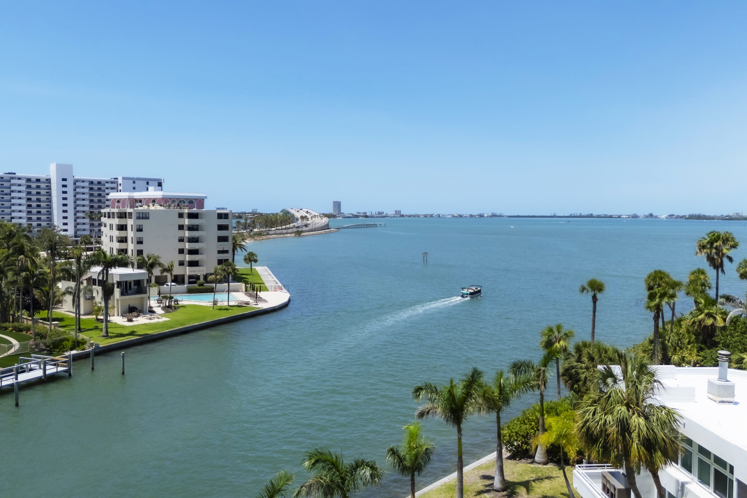 Aerial view of Sarasota Bay with high-rise buildings, palm trees, and a boat cruising on the water under a clear blue sky.