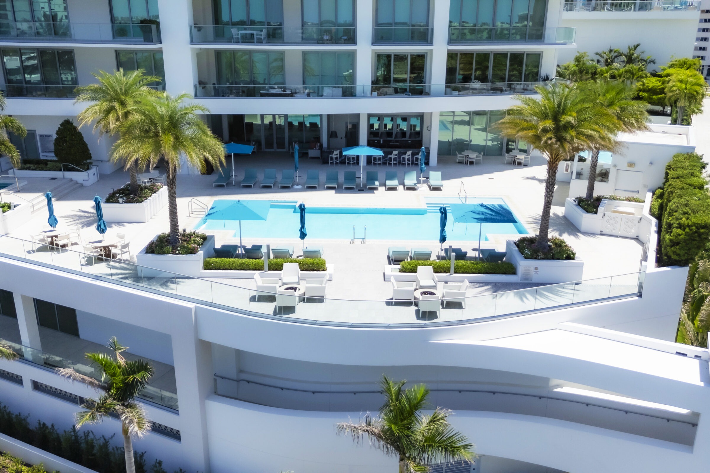 Luxurious modern building features a rooftop pool with lounge chairs, blue umbrellas, and palm trees, creating a serene outdoor retreat.