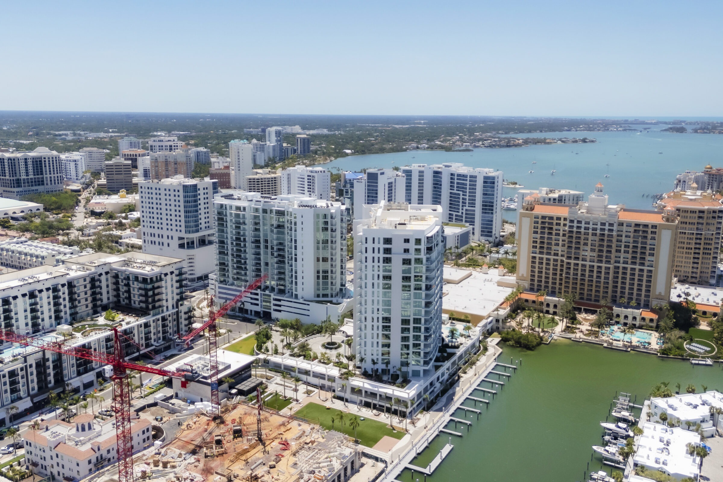 Aerial view of Sarasota, Florida, showcasing modern high-rise buildings, construction cranes, and marina, with Sarasota Bay visible in the background.