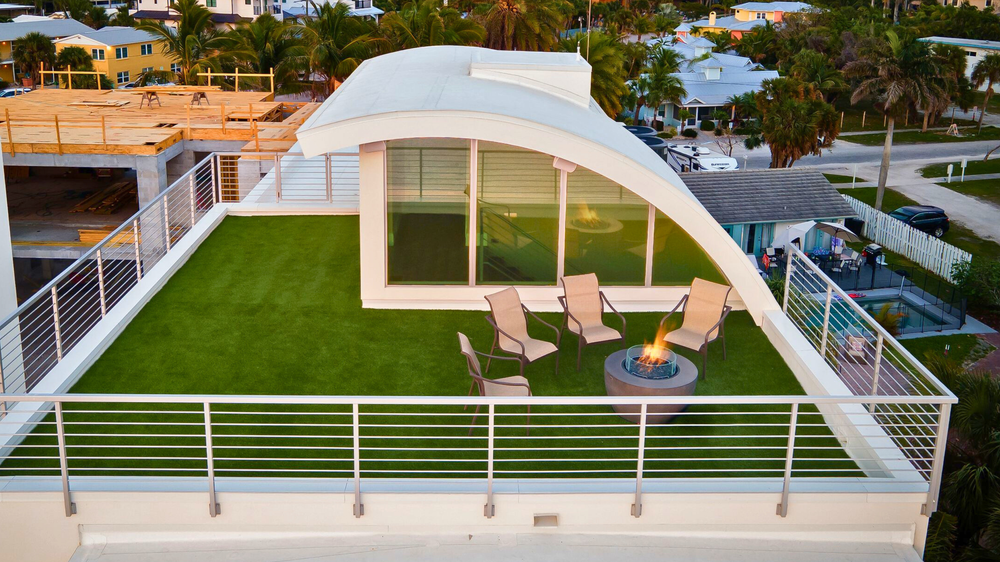 Rooftop patio with green artificial turf, modern chairs, and a fire pit. Background features tropical trees and residential neighborhood view.