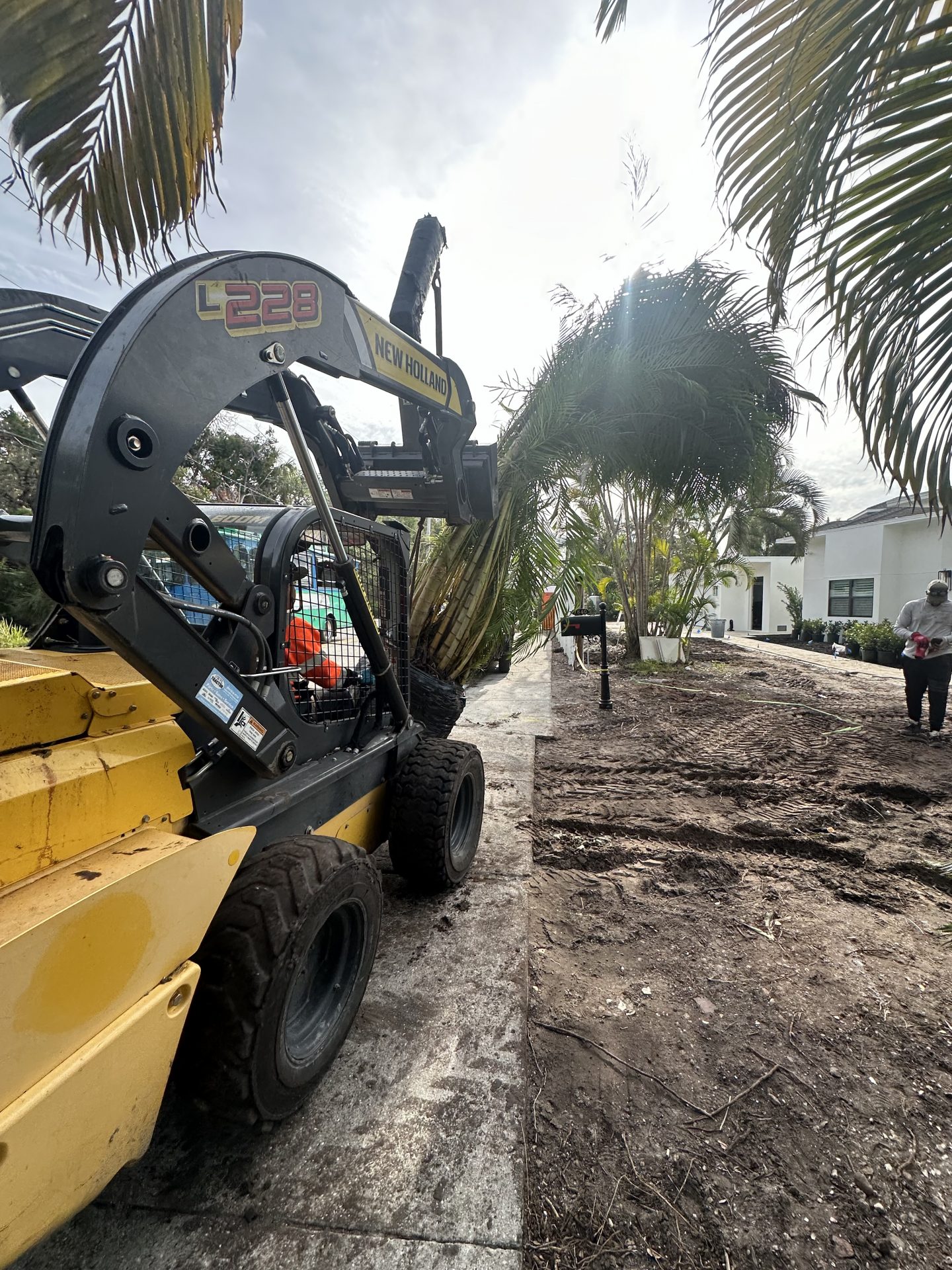 A New Holland loader operates near palm trees on a construction site. A person walks nearby, with a white building in the background.