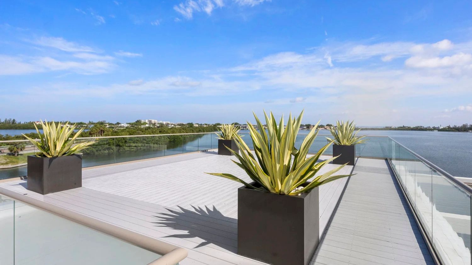 A rooftop terrace with potted plants overlooks a scenic landscape and body of water under a clear blue sky. Glass railing surrounds the deck.