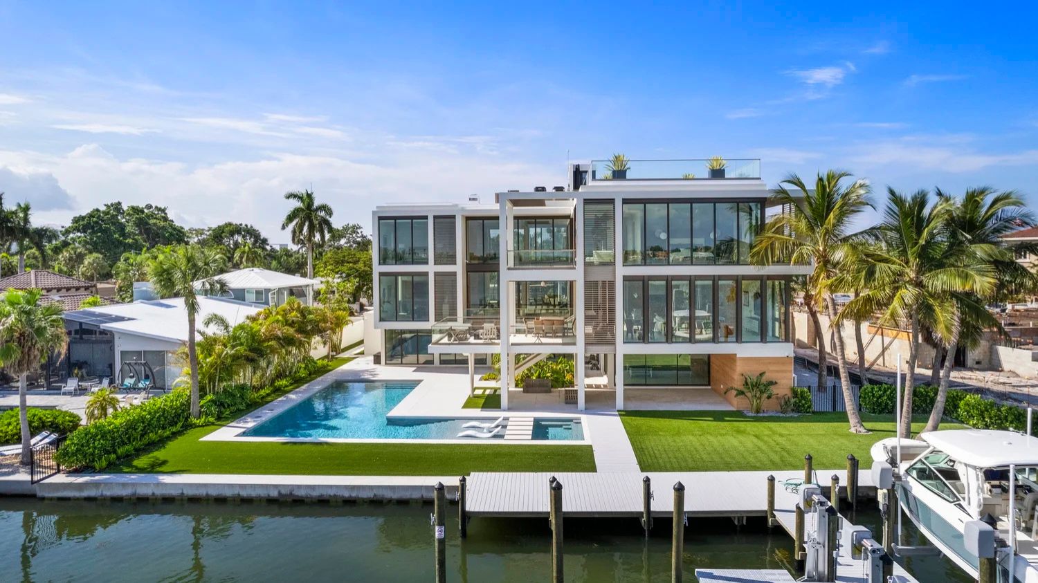 Modern waterfront home with glass façade, pool, and dock. Surrounded by lush palm trees, clear skies, and neighboring structures. No historical landmarks visible.