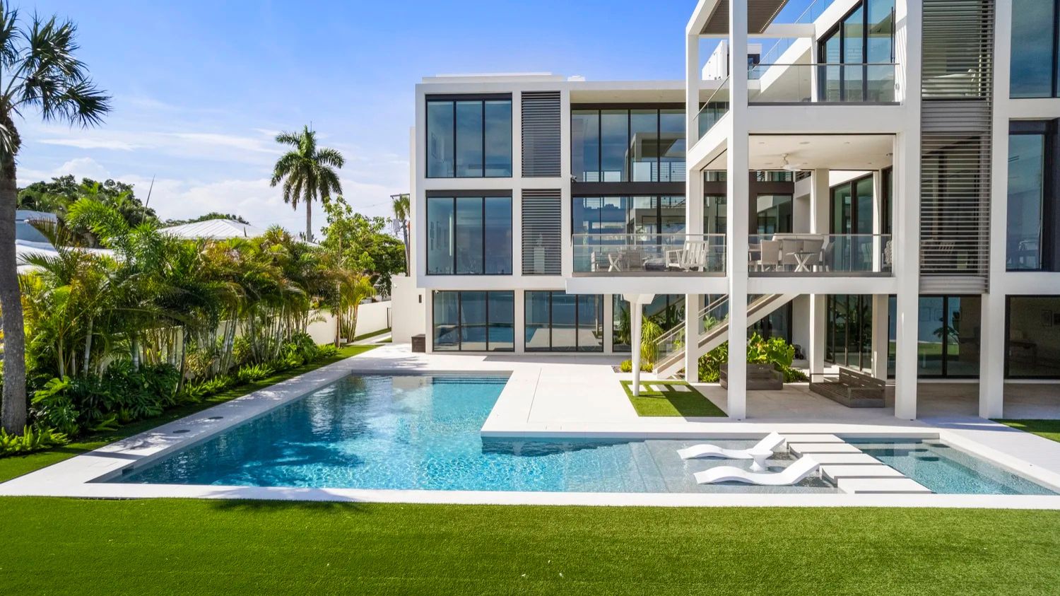 Modern white building with large windows, surrounded by palm trees. Features a spacious poolside area and well-manicured green lawn under a clear sky.