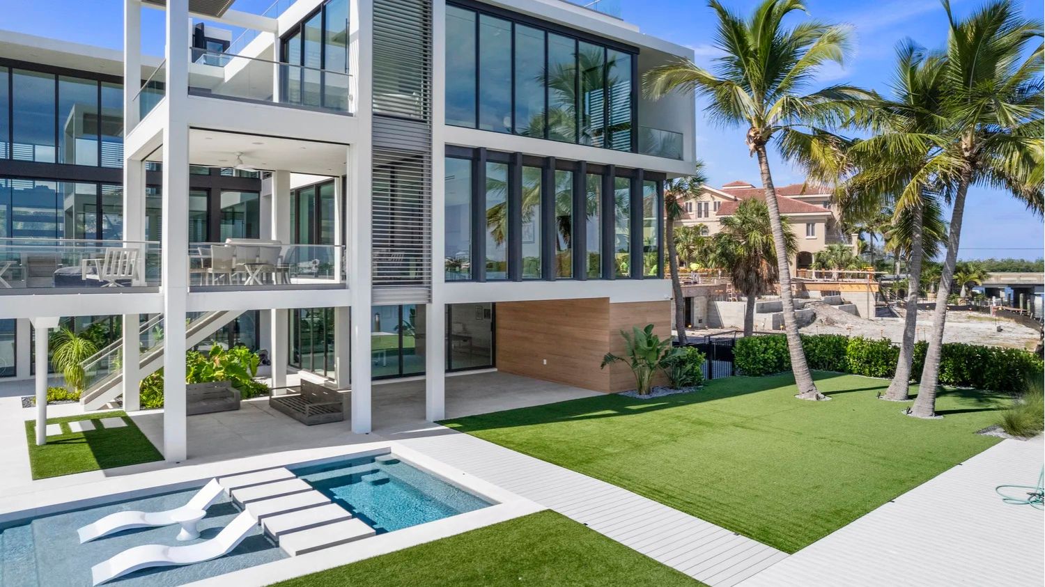 Modern house with large glass windows, pool, outdoor seating, and palm trees. Luxurious setting with neatly landscaped lawn and clear, sunny skies.