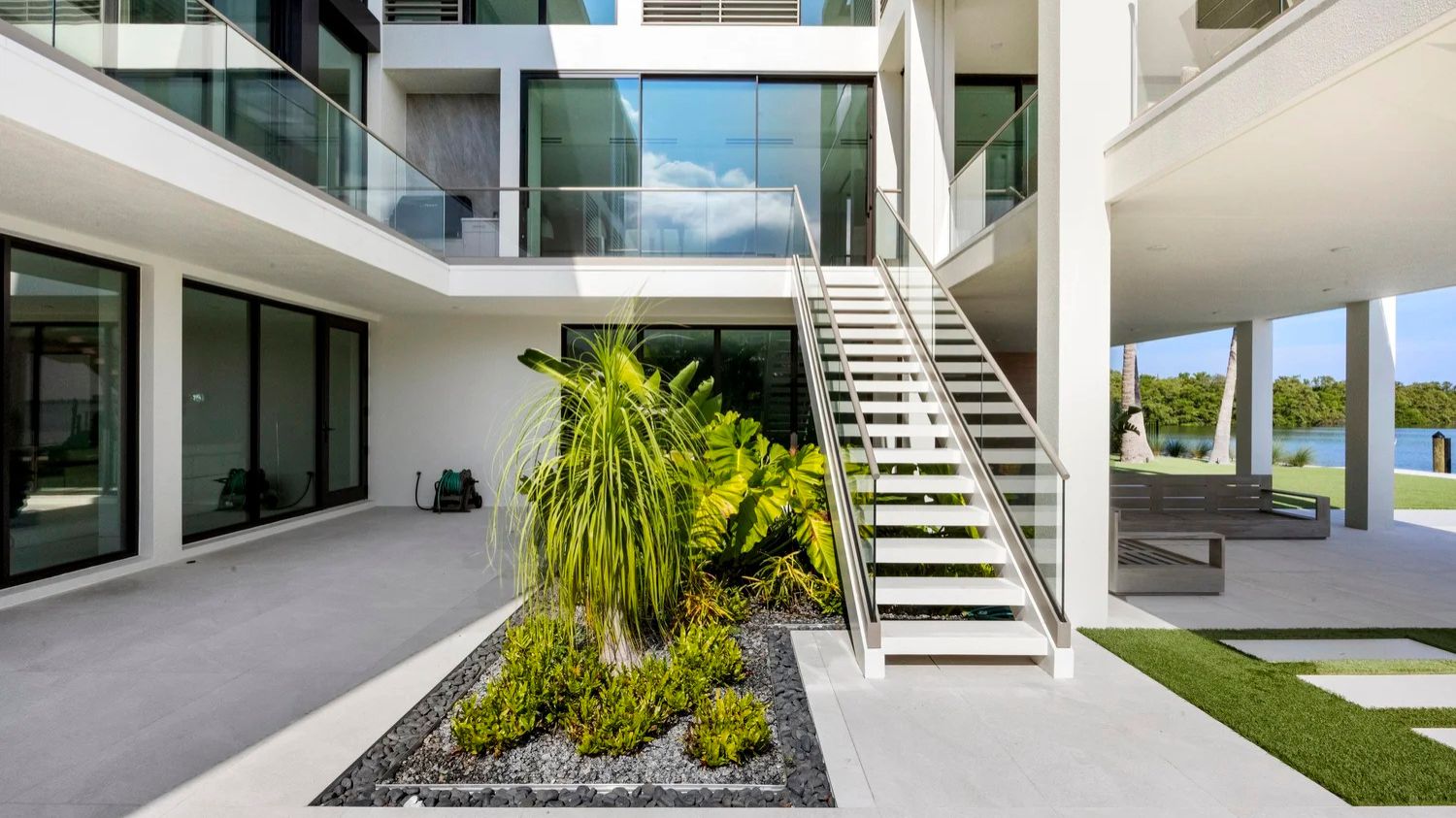Modern outdoor space featuring glass railings, white stairs, lush greenery, and a view of the water, creating a serene, architectural ambiance.