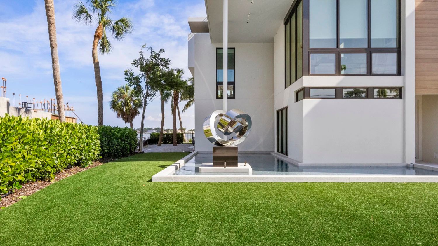 Modern house with a large metal sculpture, surrounded by palm trees and greenery, overlooks a waterfront. Sunny, blue skies enhance the scene.