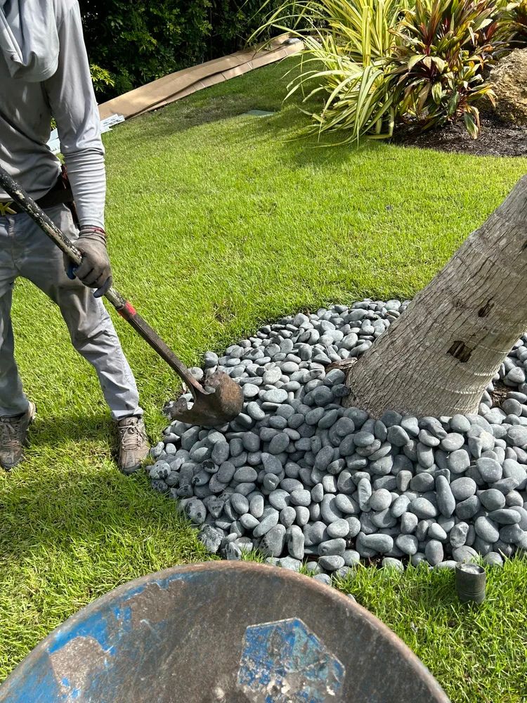 A person wearing gray uses a shovel to arrange gray stones around a tree on a grassy lawn with surrounding plants.
