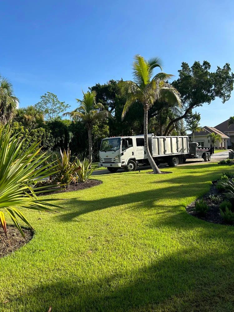 A white truck parked on a lush green lawn with palm trees and clear blue sky, near residential houses.