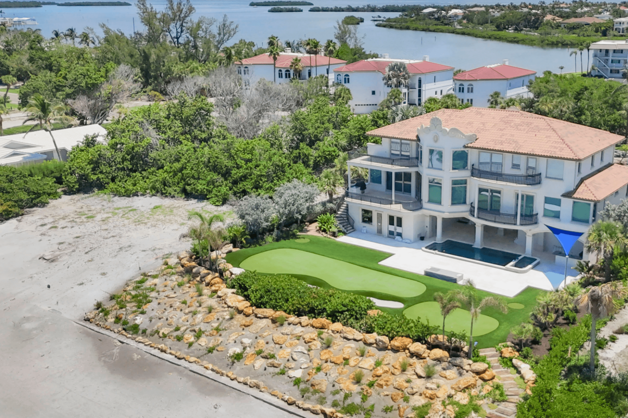 Luxurious waterfront villa with modern architecture, surrounded by lush greenery, a private pool, and putting green, overlooking a scenic body of water.