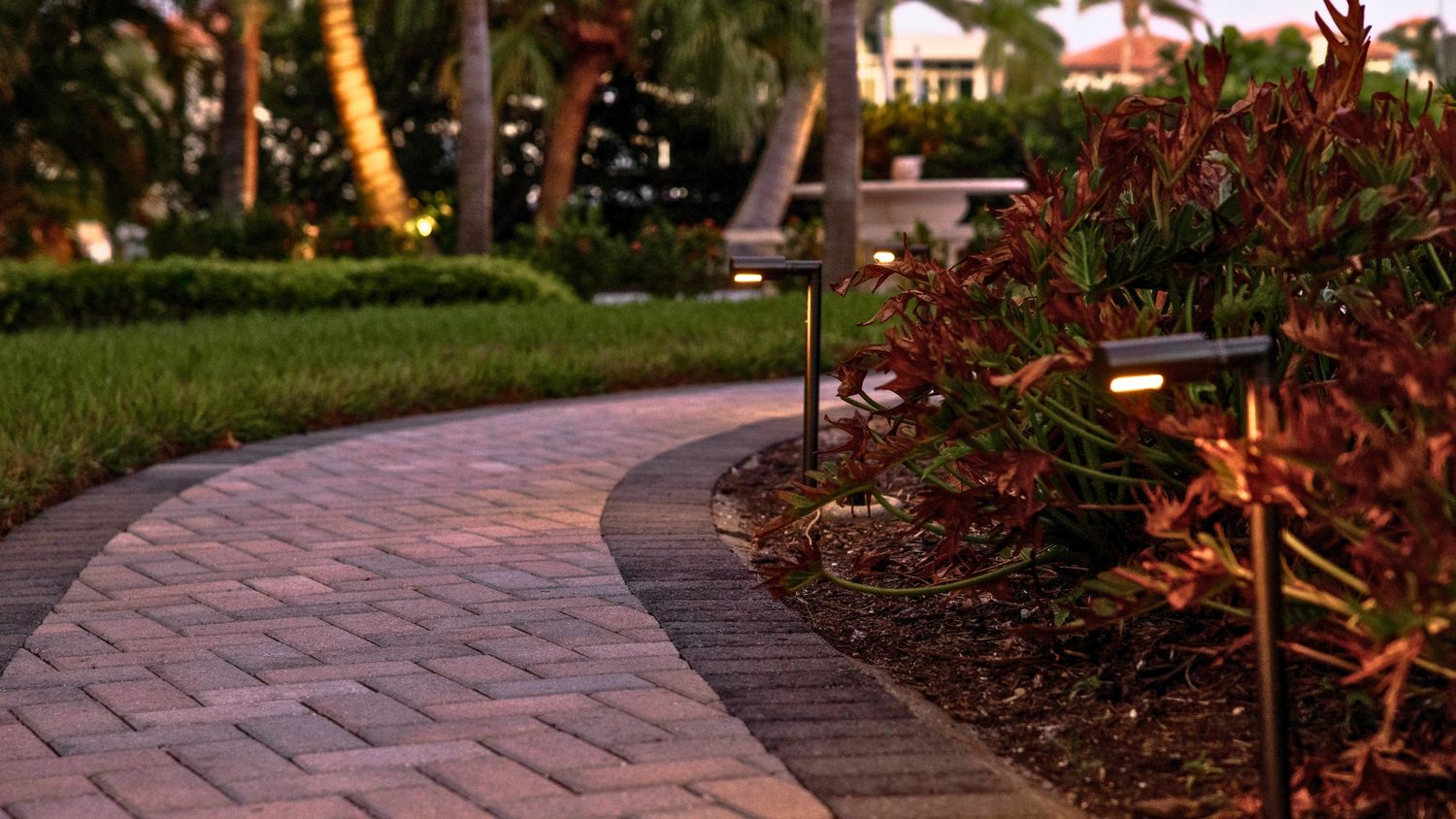 Curved brick path with small lights and lush greenery, leading through a garden setting at dusk. Palms and shrubs enhance serene ambiance.