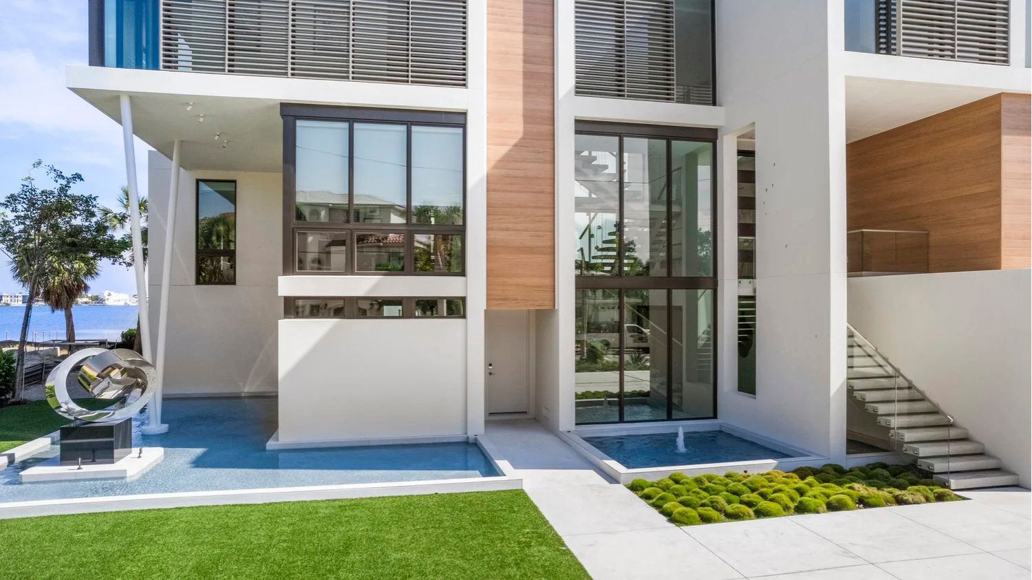 Modern building exterior with large windows, white walls, and a metal sculpture by a water feature. Green lawn and nearby water visible.