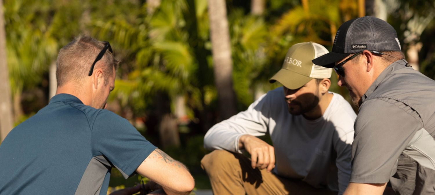 Three people outdoors, wearing caps and sunglasses, engaged in a focused discussion surrounded by tropical greenery, with sunlight creating natural highlights.