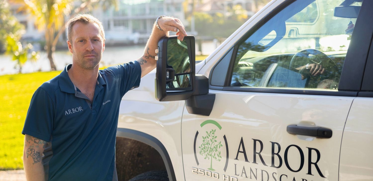 A person in an "ARBOR" shirt stands by a branded truck, with a sunny park background and waterway view.