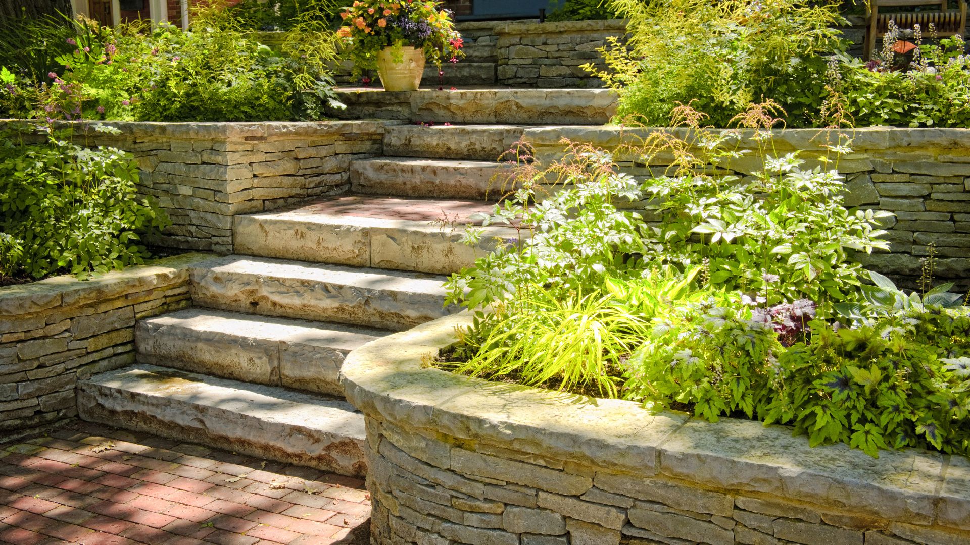 Stone terraced garden with lush green plants and vibrant flowers, featuring curving stone steps leading upward in sunlit outdoor setting.