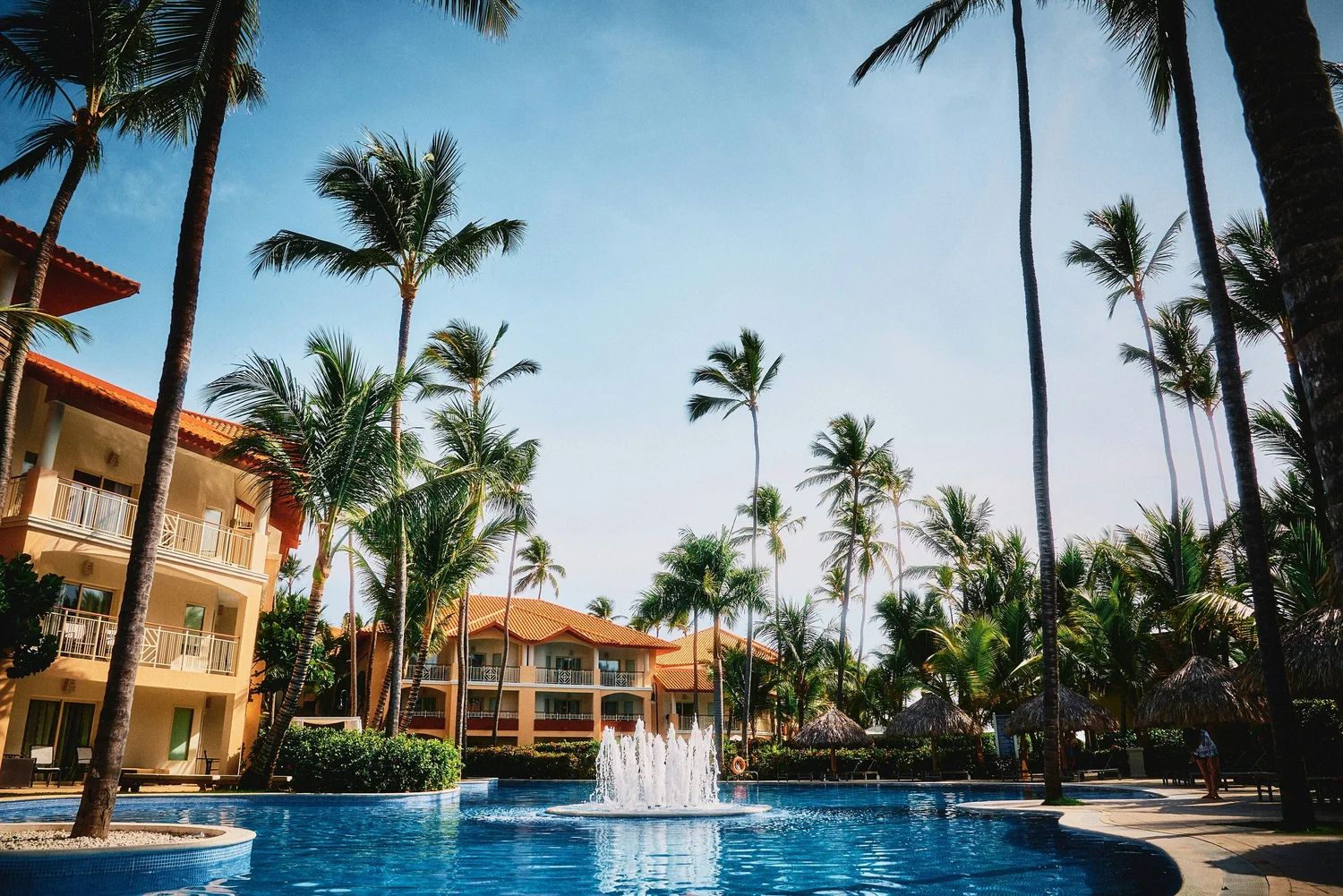 A tropical resort with palm trees, a fountain in a blue pool, surrounded by beige buildings and thatched umbrellas under a clear sky.
