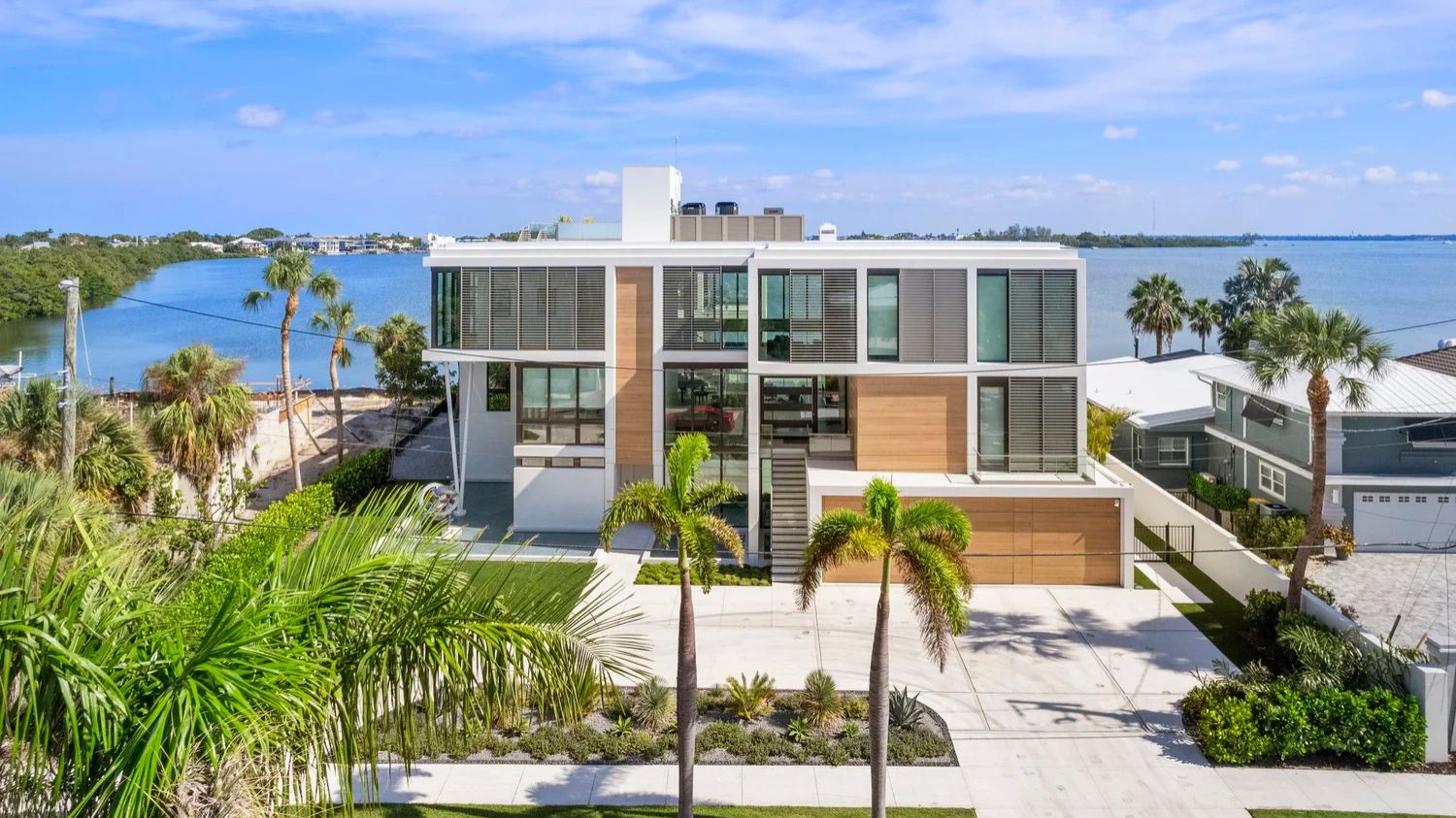 Modern waterfront house with large windows, surrounded by palm trees and overlooking a serene bay. Clear blue sky and lush greenery enhance the scene.