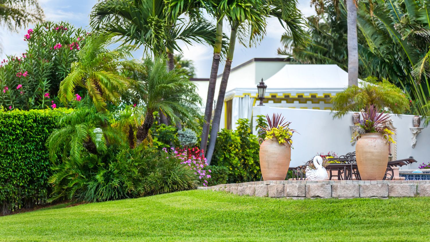 Lush garden with palm trees, vibrant flowers, large clay pots, and a swan sculpture on a stone platform, surrounded by manicured greenery.