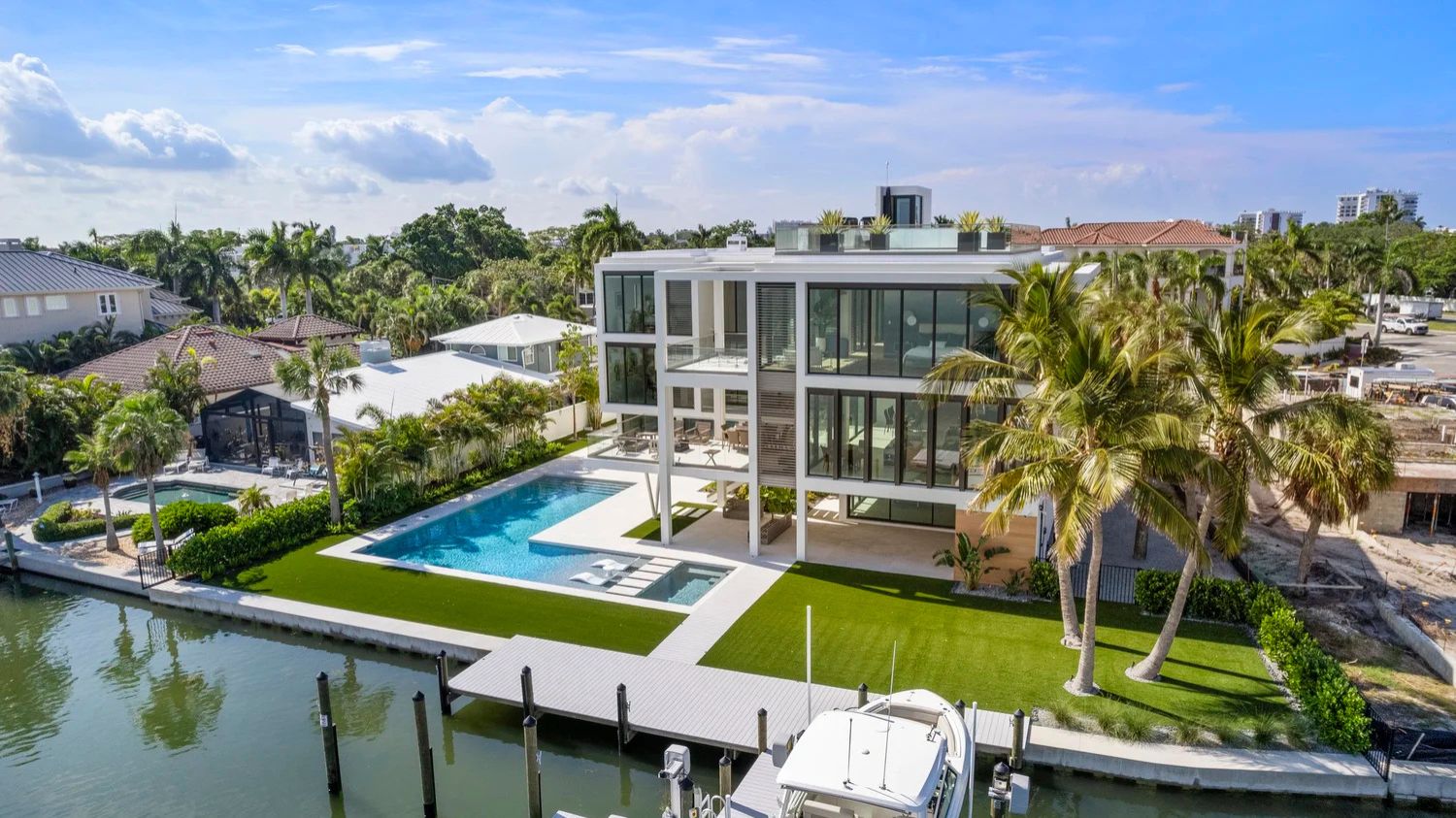Modern waterfront house with glass facade, pool, and palm trees. Surrounded by greenery, docked boat, and neighboring homes under a clear blue sky.